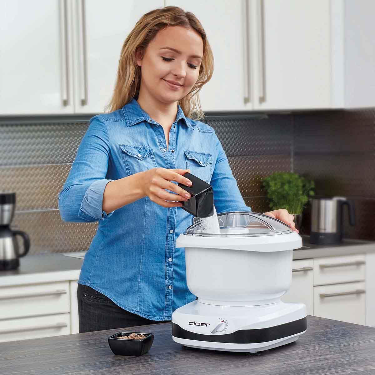 Frau gibt Zutaten in einen weißen Küchenmixer. Sie steht in einer Küche.
