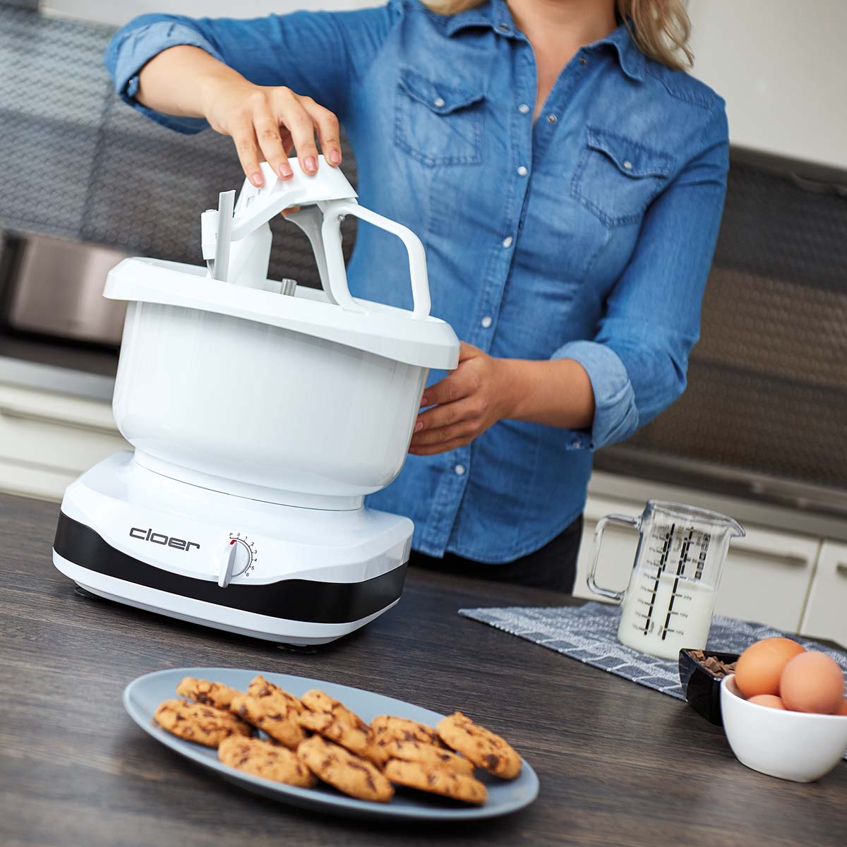 Eine Person in einem blauen Jeanshemd benutzt eine weiße Küchenmaschine. Kekse, Eier und Milch stehen auf der Theke.
