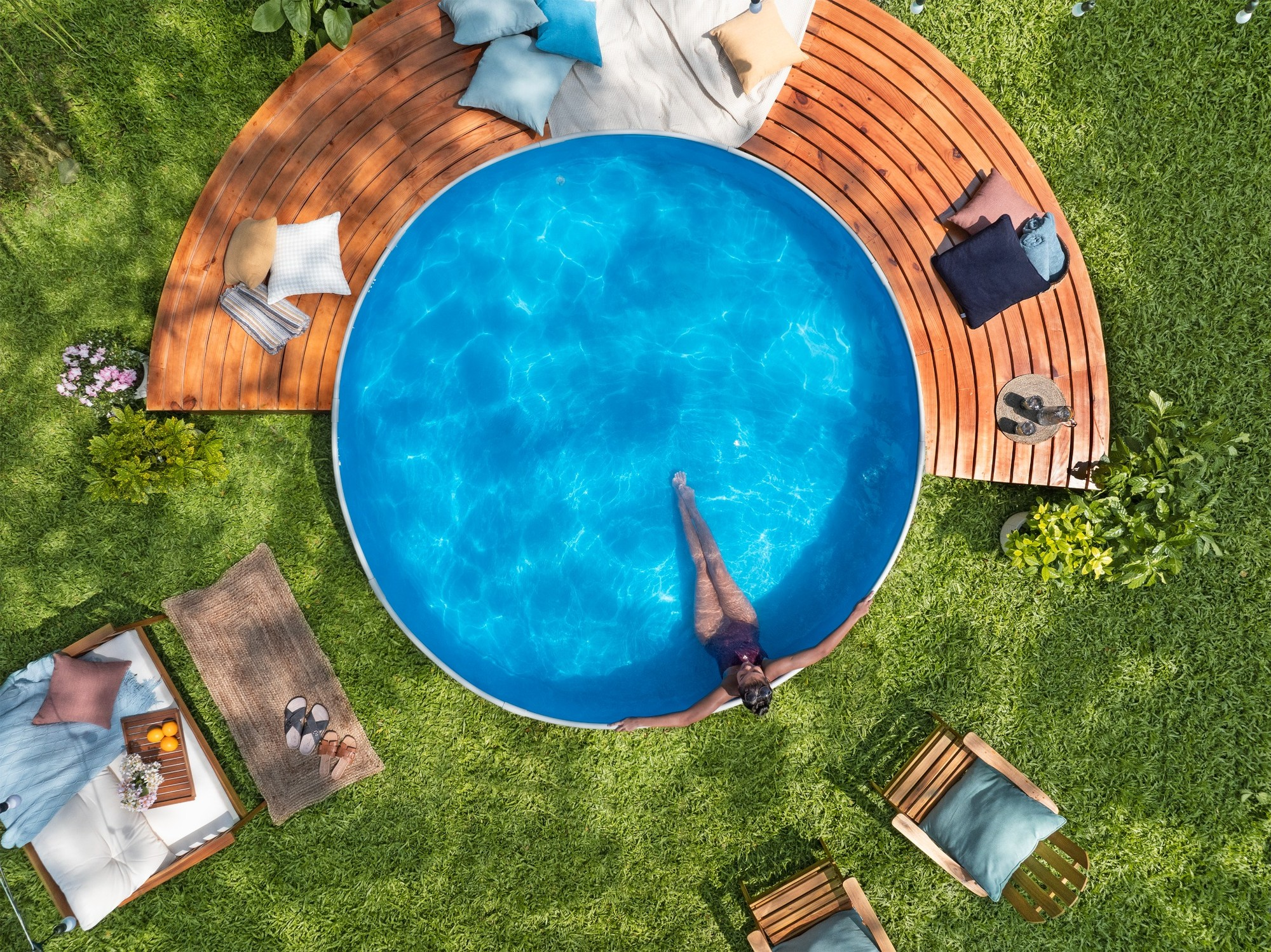 Eine Frau entspannt im Pool auf einer Holzterrasse mit Liegestühlen und Kissen im Garten.