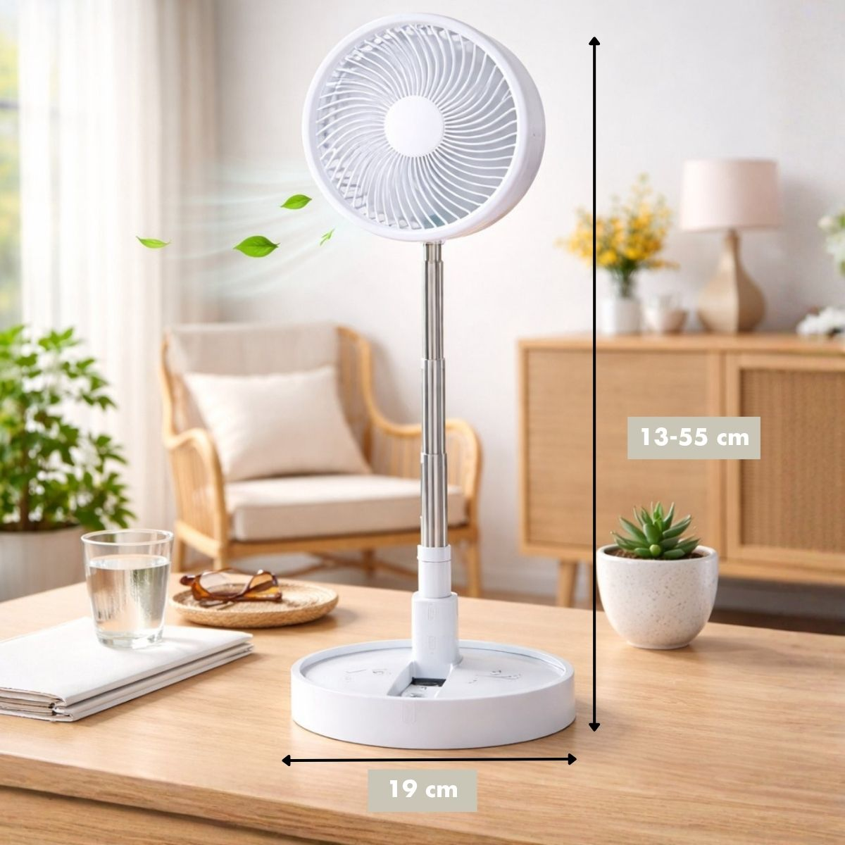 Un ventilateur blanc et pliable se dresse sur une table en bois. Des feuilles vertes volent.