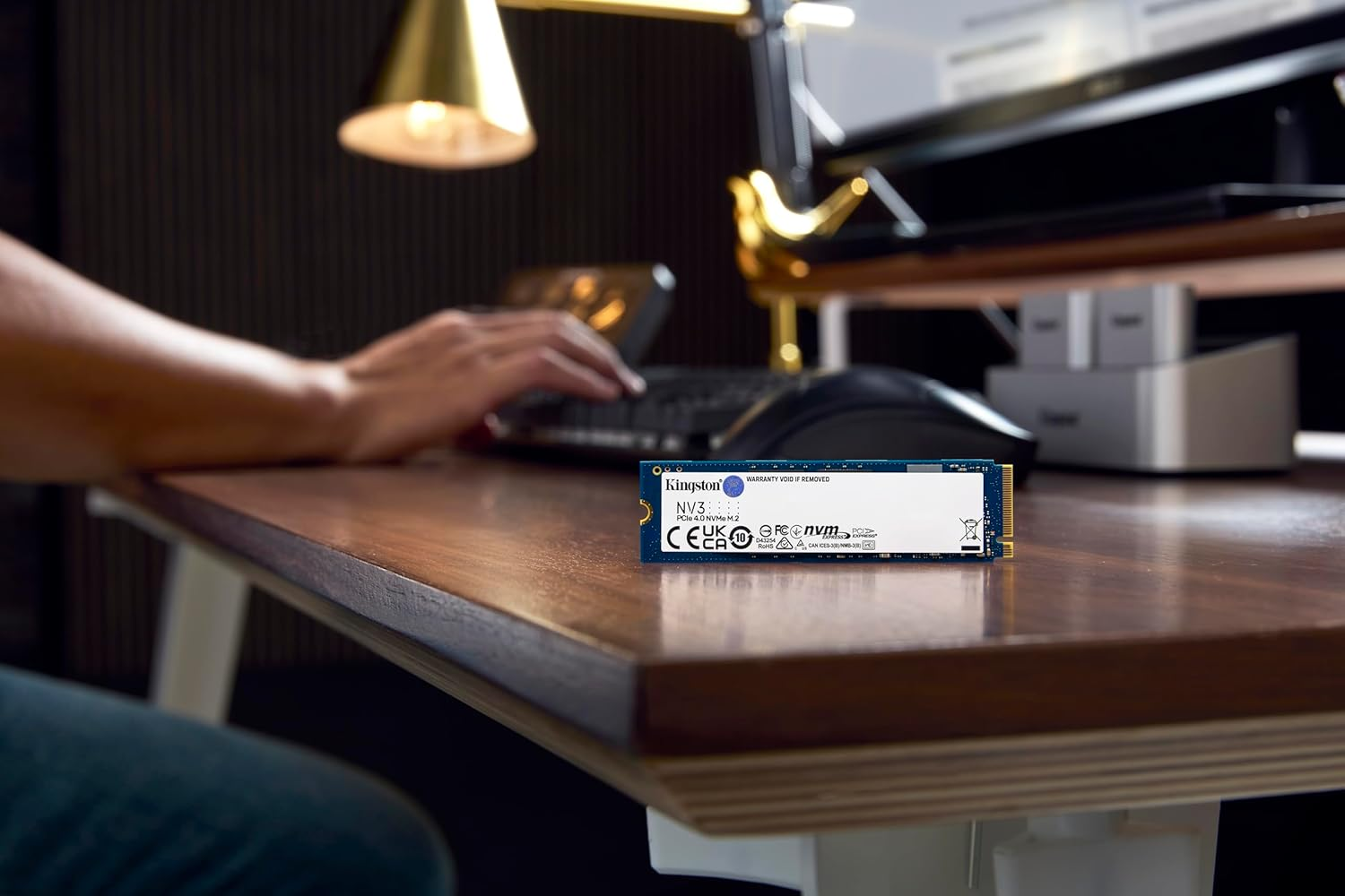 Eine Kingston NVMe M.2 SSD liegt auf einem Holztisch, im Hintergrund tippt eine Person auf einer Tastatur.
