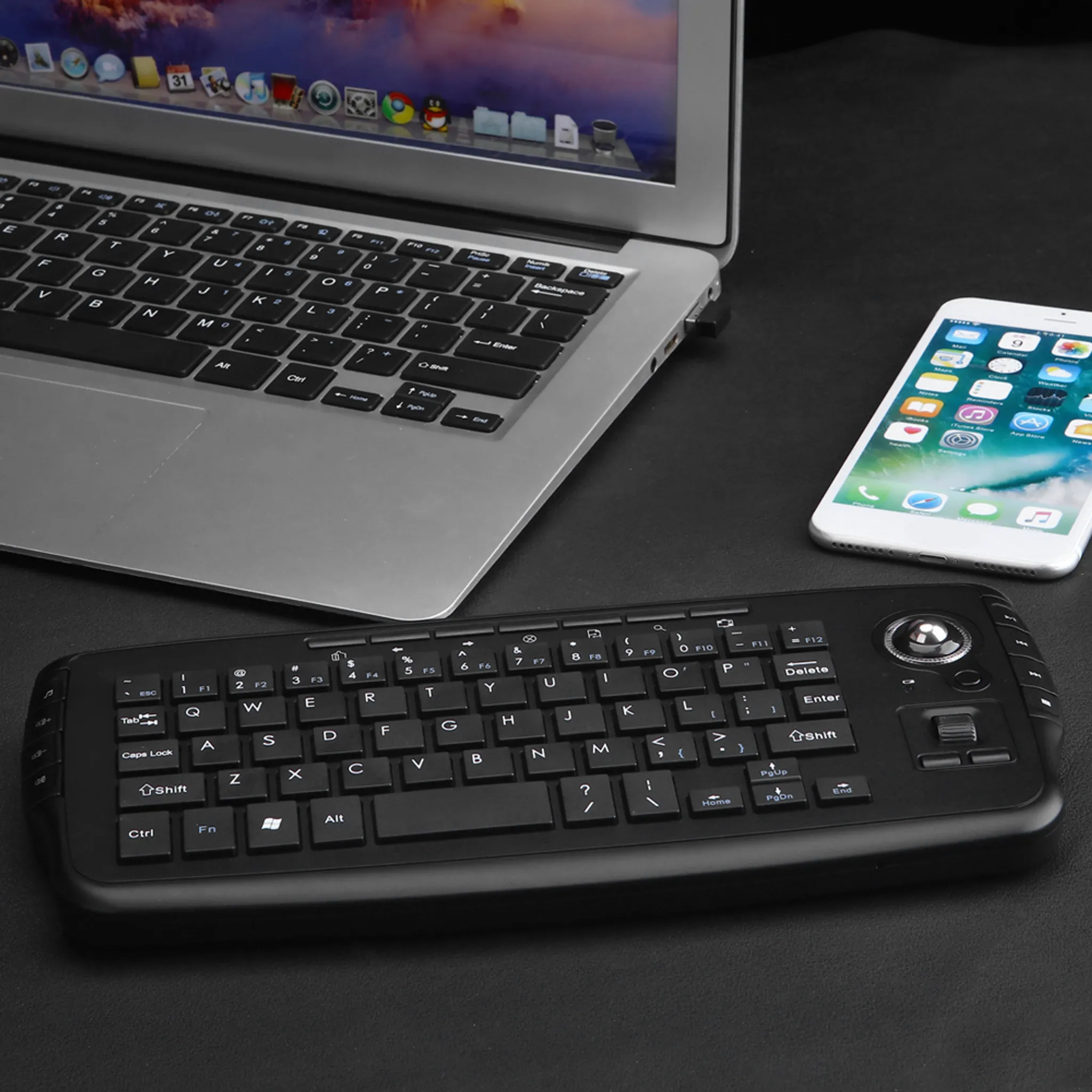 Ein Laptop, Smartphone und eine kabellose Tastatur mit Trackball liegen auf dunkler Fläche.