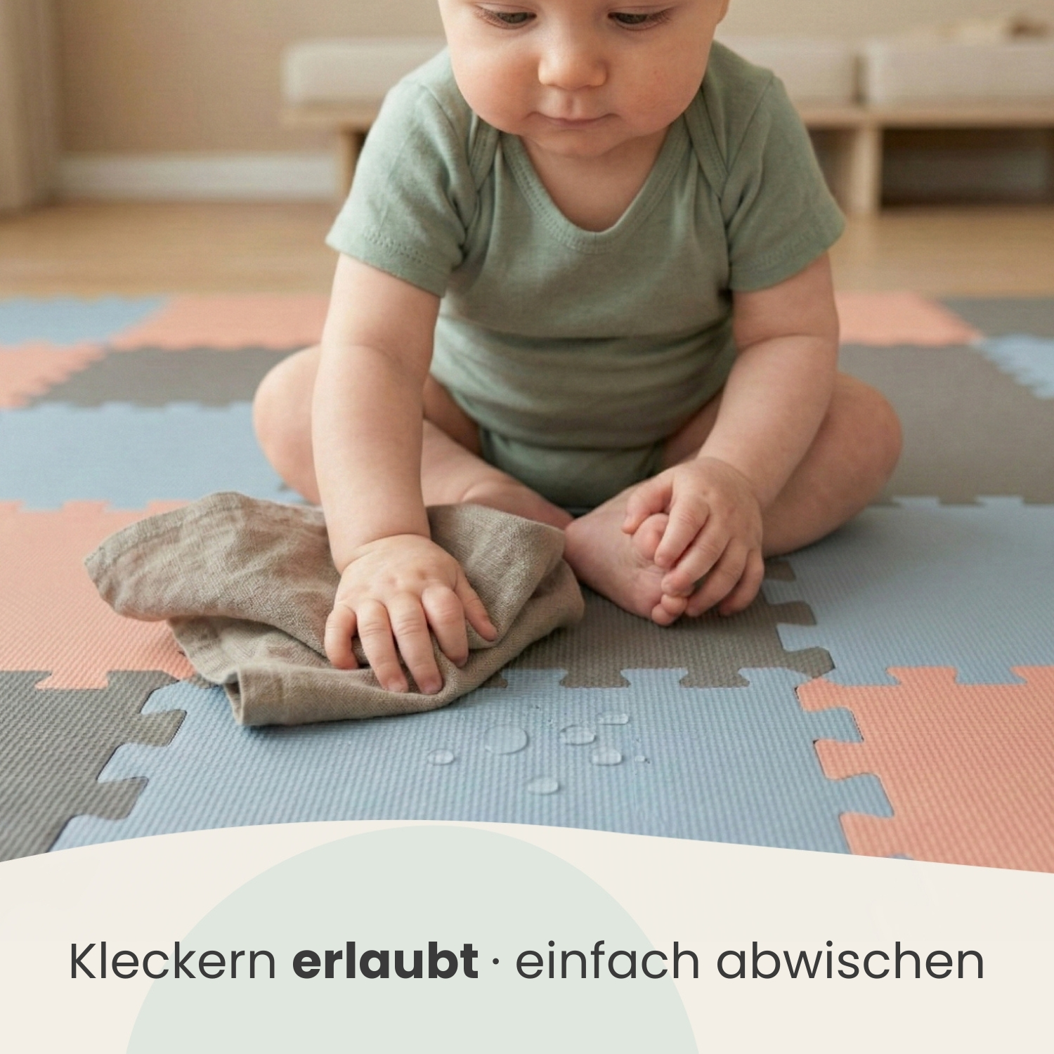 Ein Baby in einem grünen Body sitzt auf einer bunten Spielmatte und wischt Wasser auf.