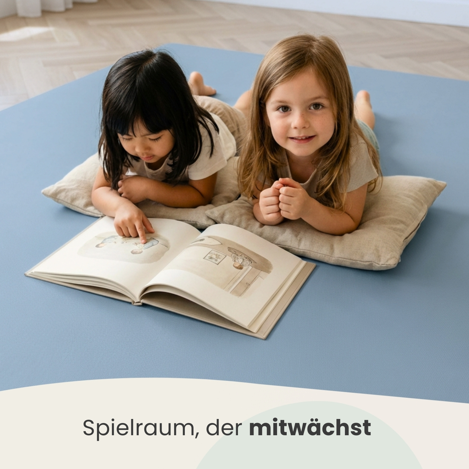 Zwei Mädchen liegen auf einem blauen Teppich und lesen ein Buch, eines zeigt darauf.