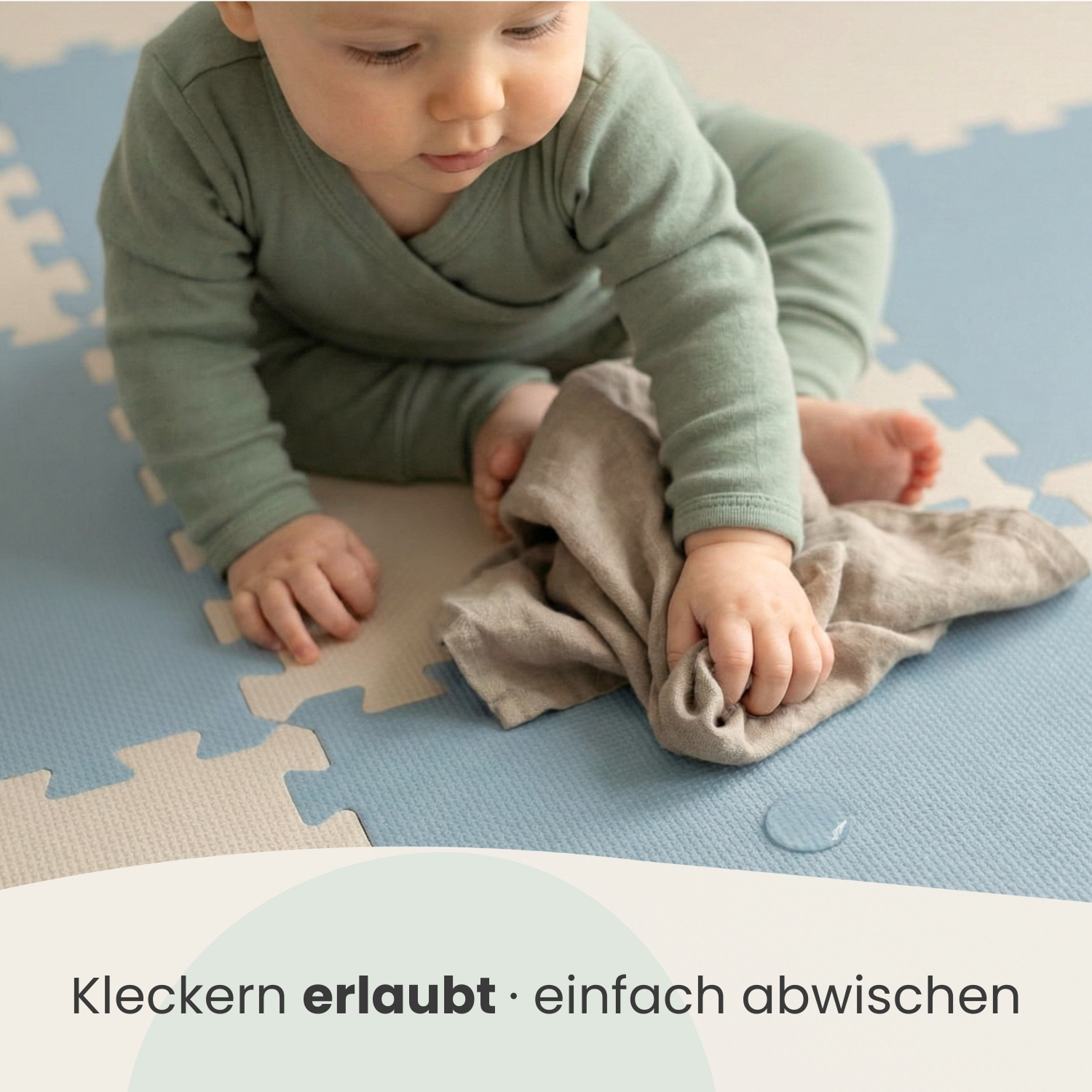 Ein Baby in einem grünen Strampler wischt mit einem Tuch über eine blaue Matte.
