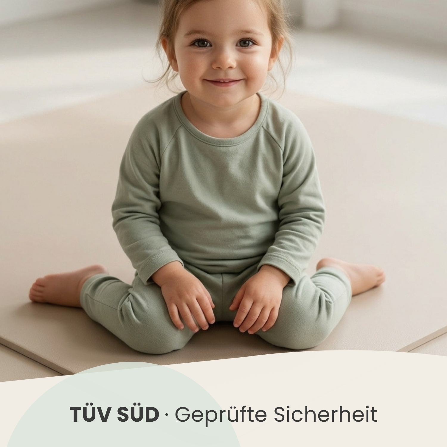 Kleines Kind in grüner Kleidung sitzt auf einer Matte und lächelt.