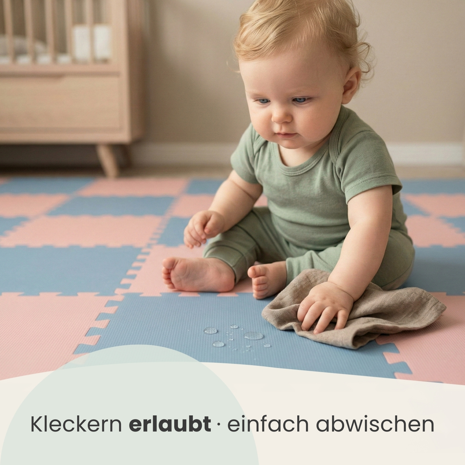 Baby in grüner Kleidung sitzt auf einer rosa-blauen Matte und wischt verschüttetes mit einem Tuch ab.