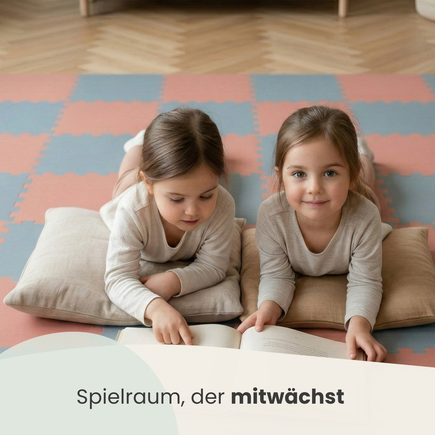 Zwei kleine Mädchen liegen auf Kissen und lesen ein Buch auf einer bunten Matte.