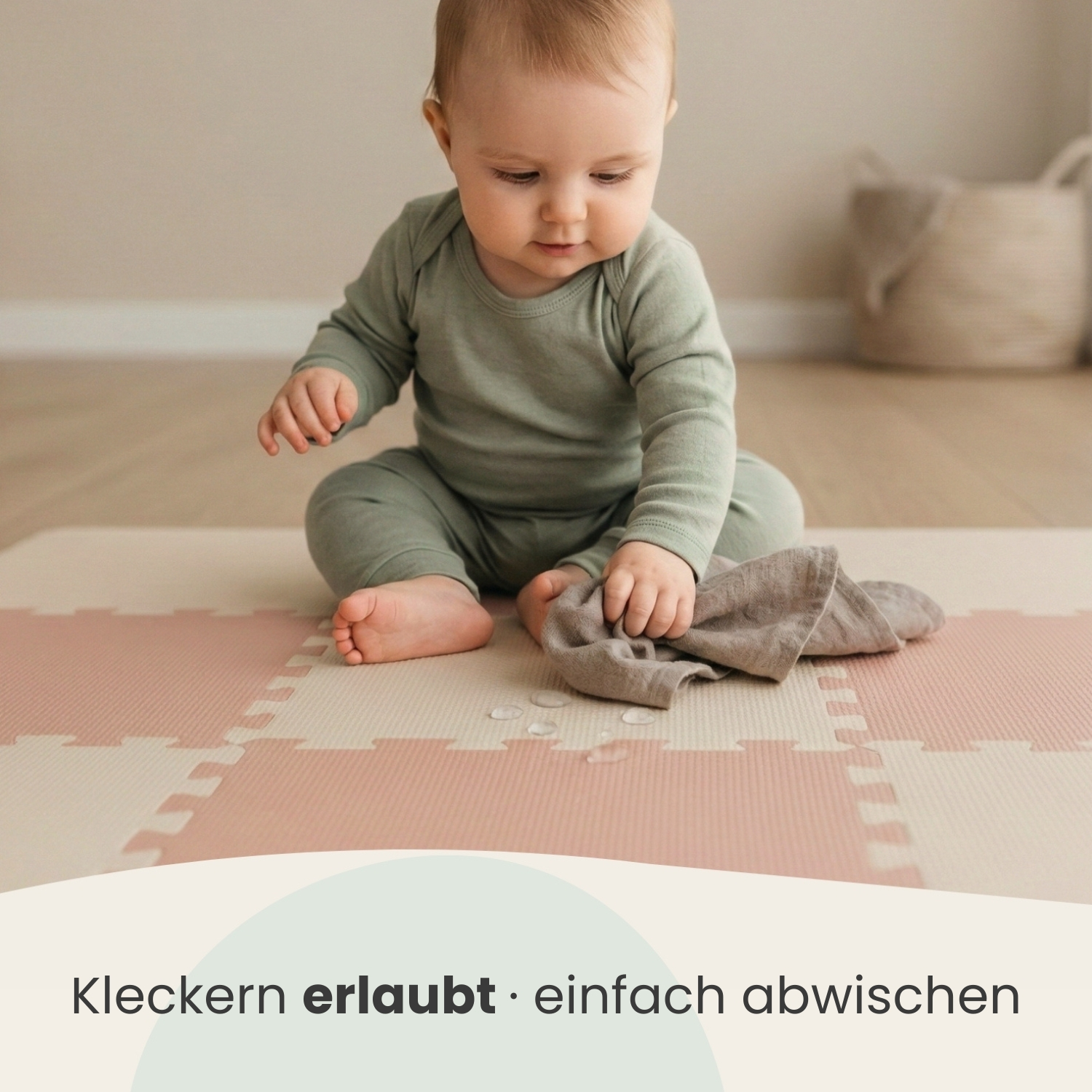 Baby in grünem Strampler sitzt auf Spielmatte. Hält Stoff. Tropfen auf Matte.