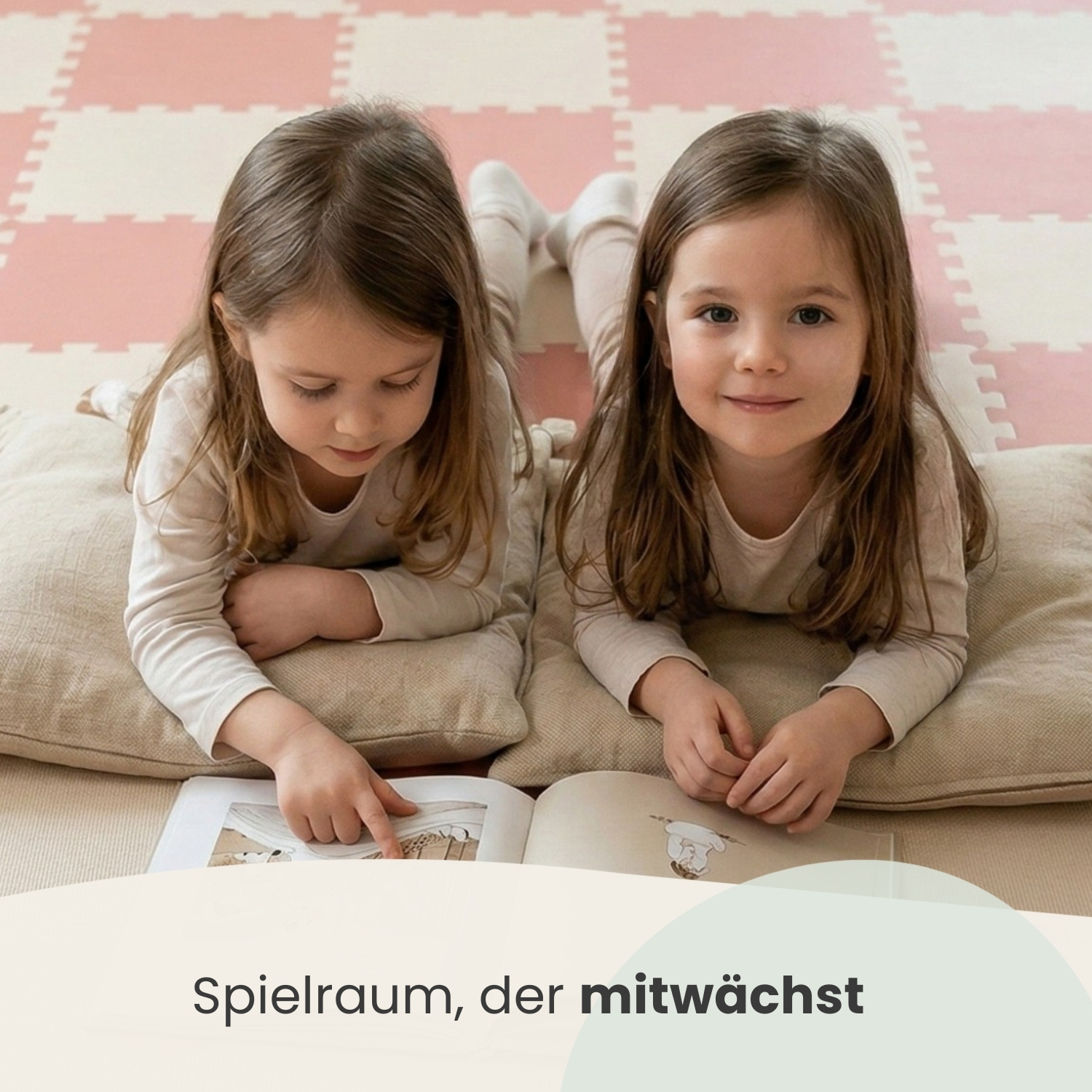 Zwei kleine Mädchen liegen auf einer Spielmatte, eines liest ein Buch, das andere schaut auf.