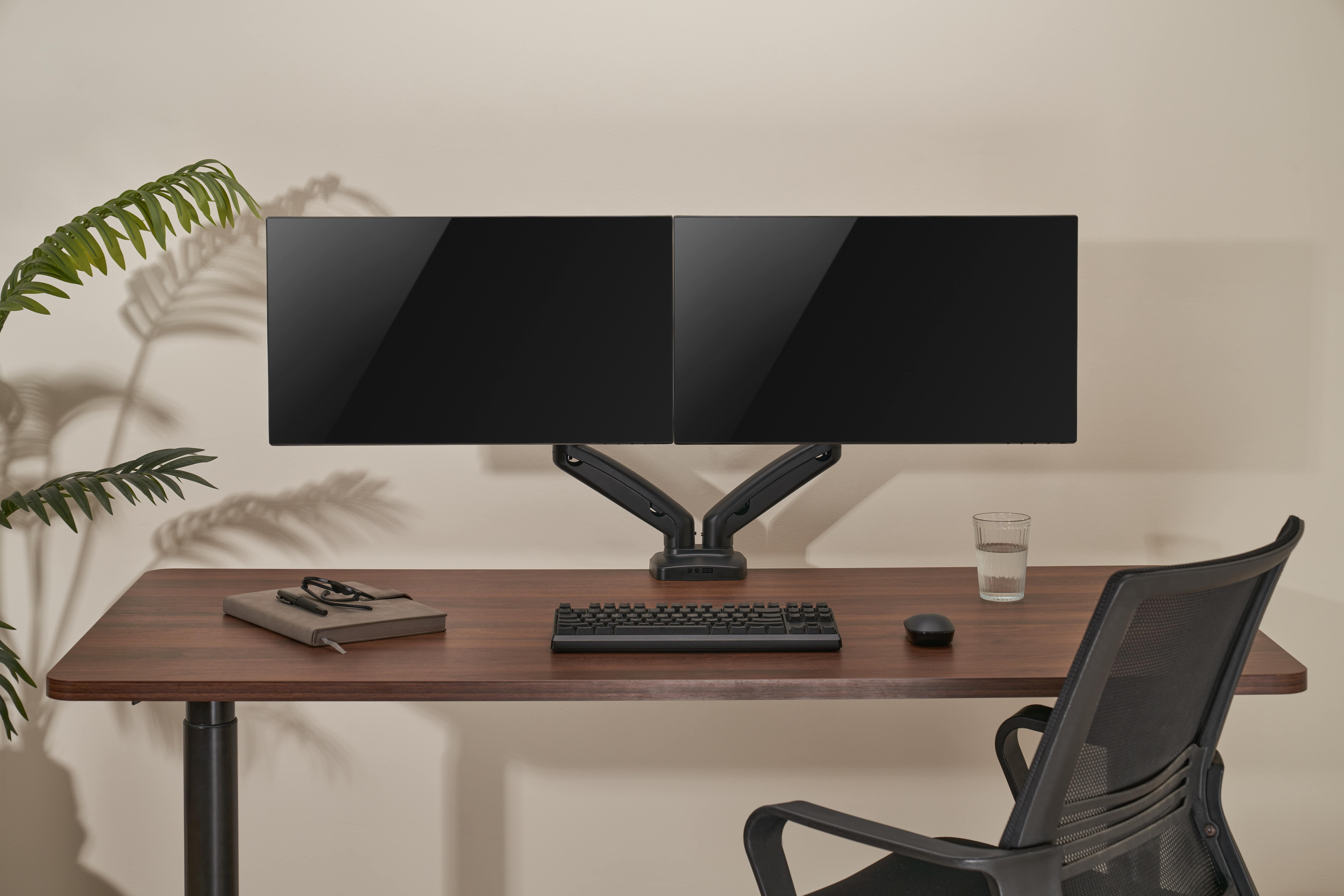 Ein Schreibtisch mit zwei Monitoren, Tastatur, Maus, Notizbuch und Glas Wasser.
