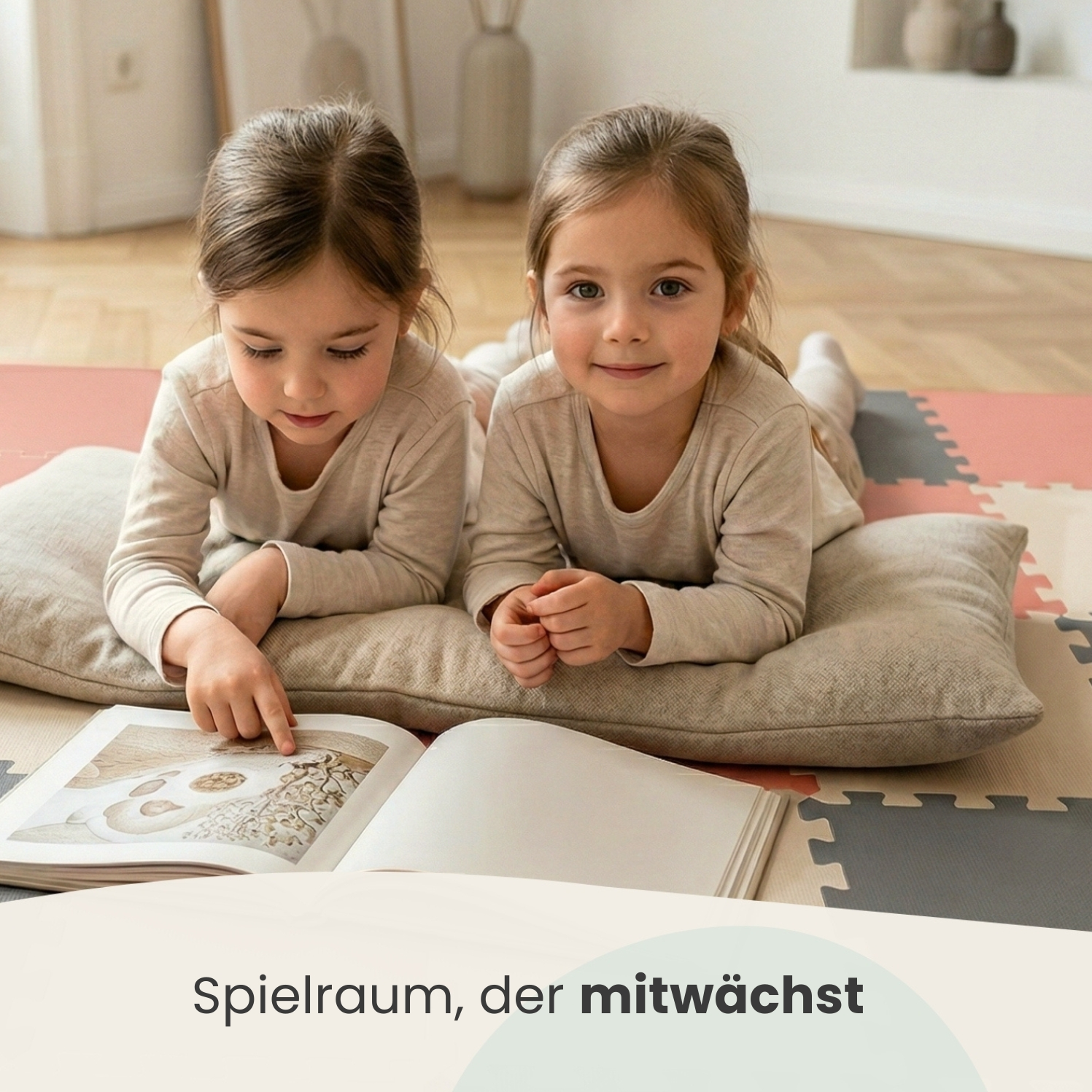 Zwei kleine Mädchen liegen auf einer Bodenmatte und lesen ein Buch. Eines zeigt auf eine Seite.