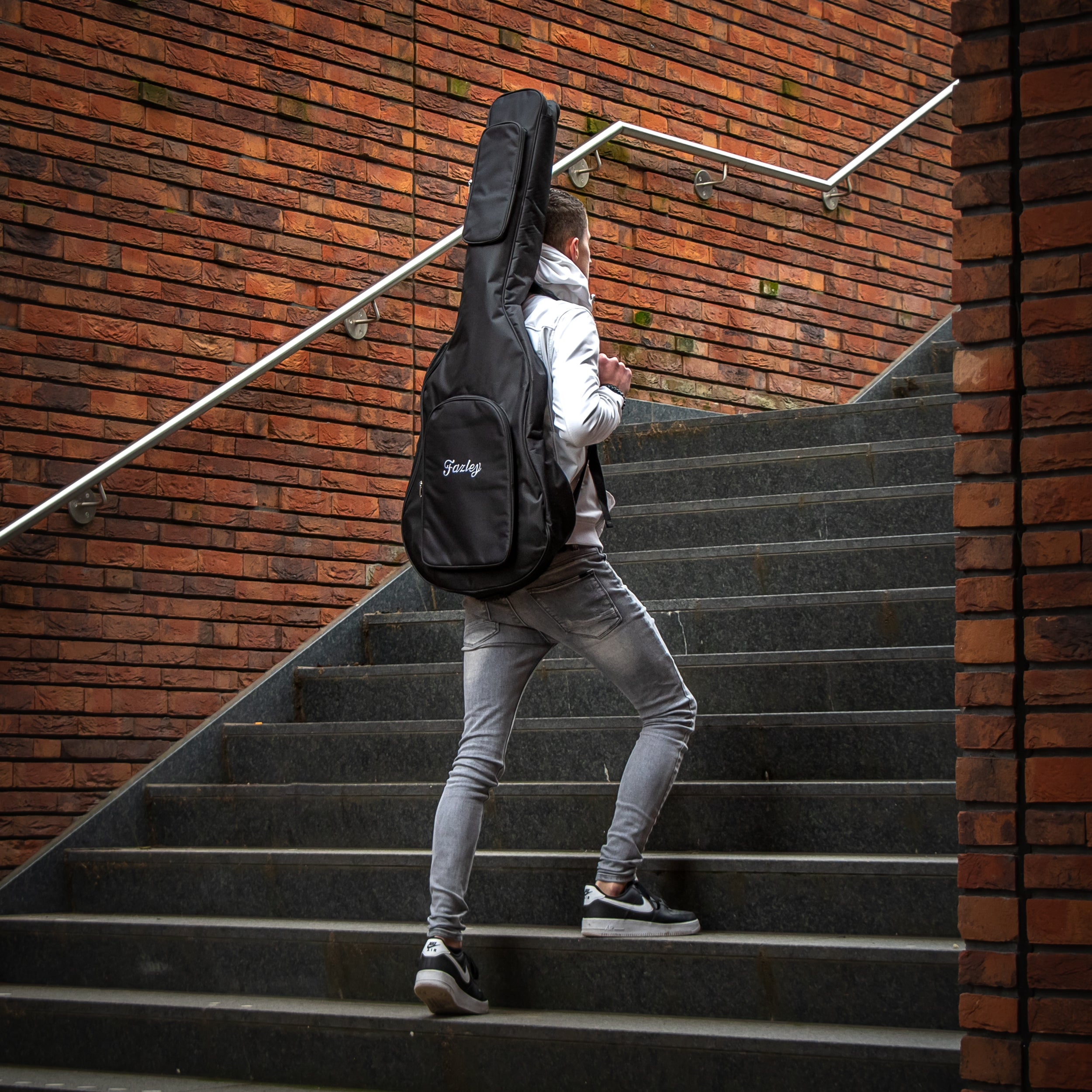 Jongere met gitaarkoffer loopt stenen trap op, rode bakstenen muur.