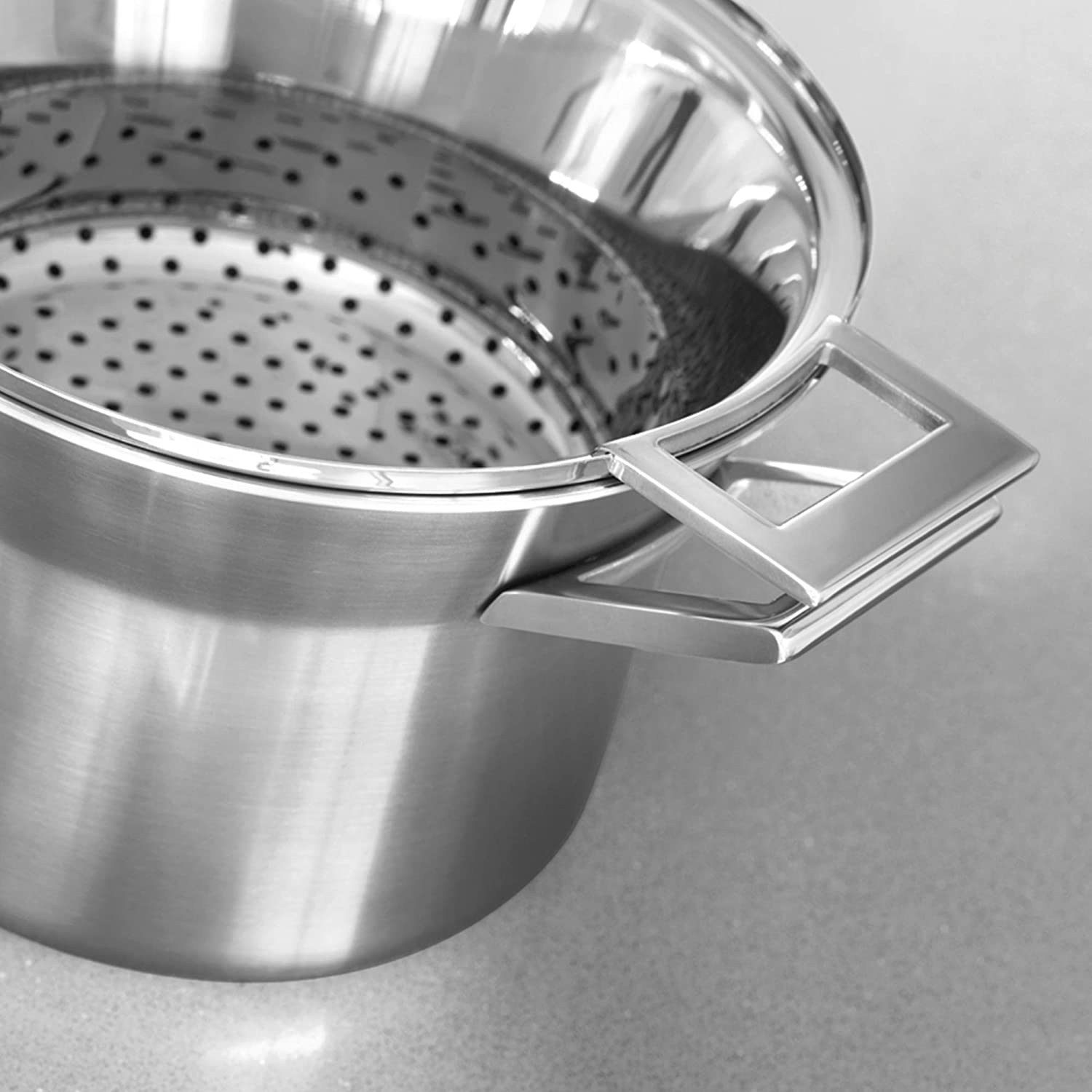 Srebrny kosz do gotowania na parze z uchwytami spoczywa na szarej powierzchni.