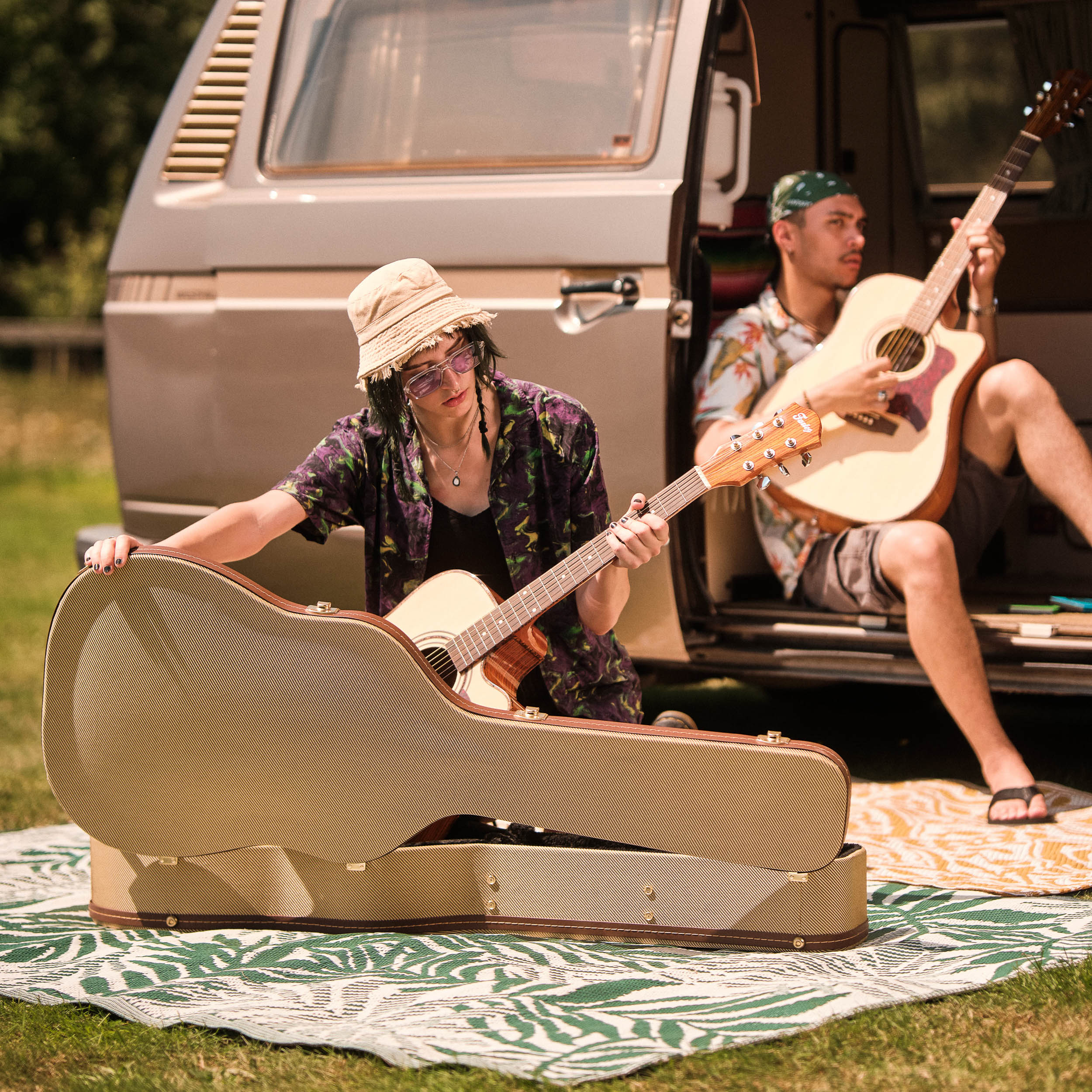 Twee mensen spelen gitaar naast een busje op picknickkleden buiten.