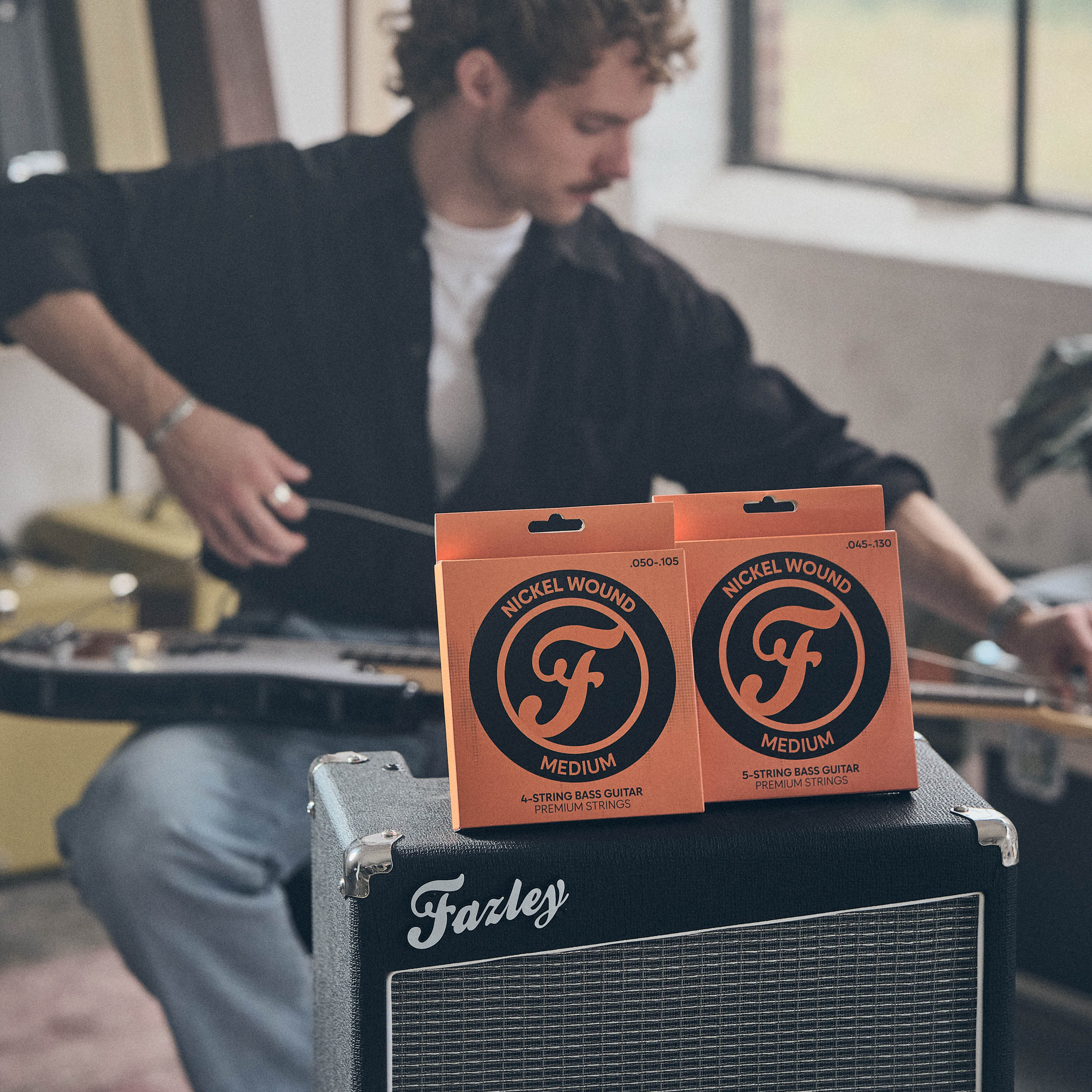 Een man bespeelt een gitaar. Twee pakken snaren rusten op een versterker.