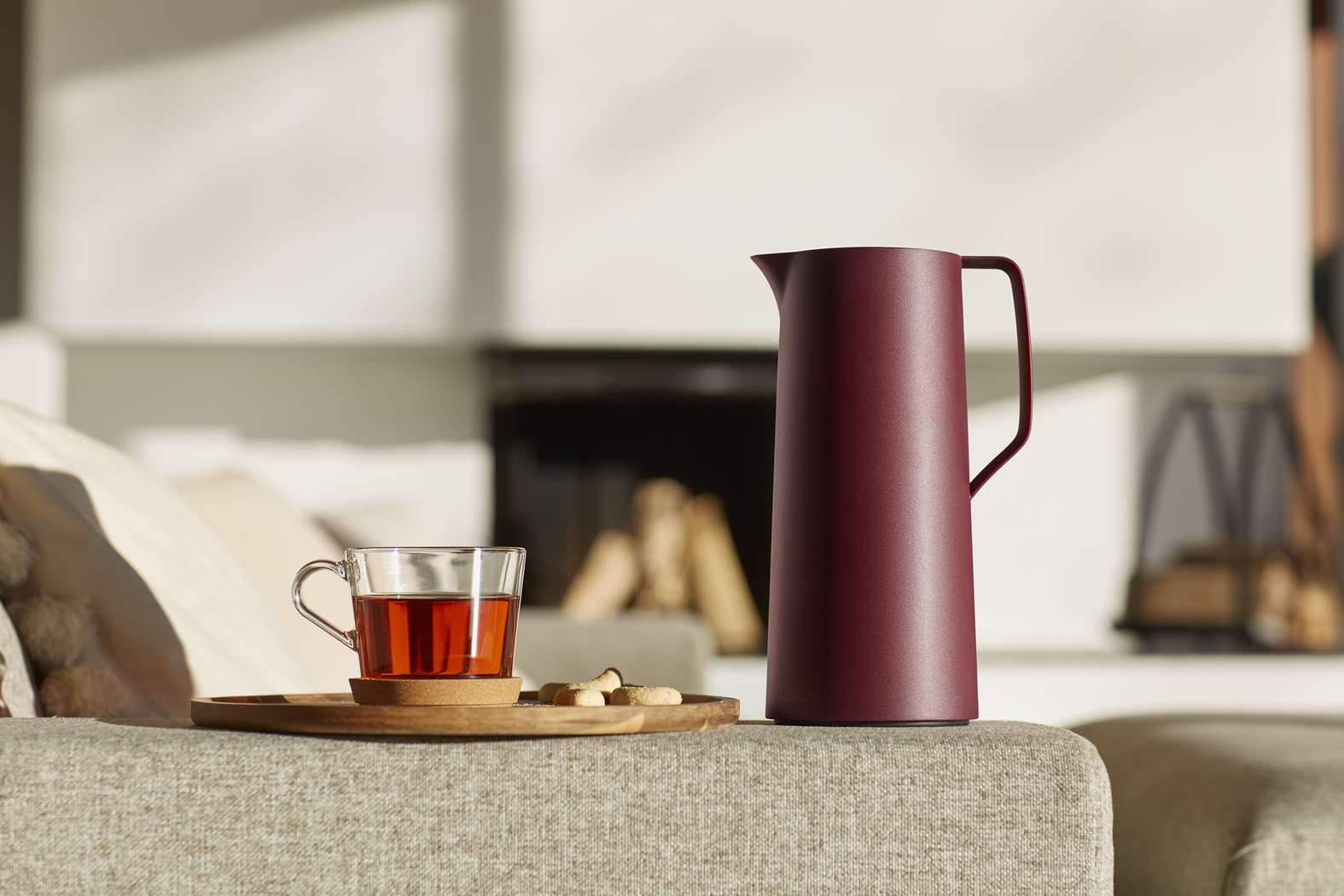 Una taza de té de cristal y un termo rojo sobre un sofá, con una chimenea al fondo.