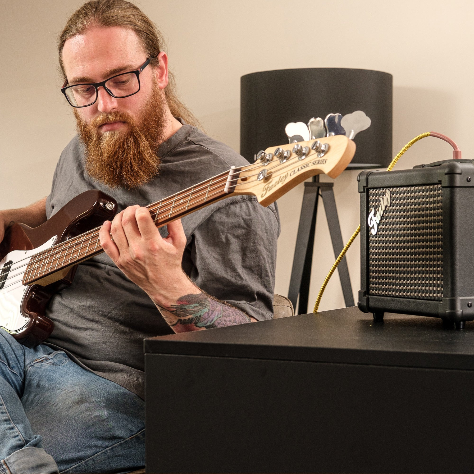 Een man met bril en baard speelt basgitaar, een kleine versterker staat ernaast.