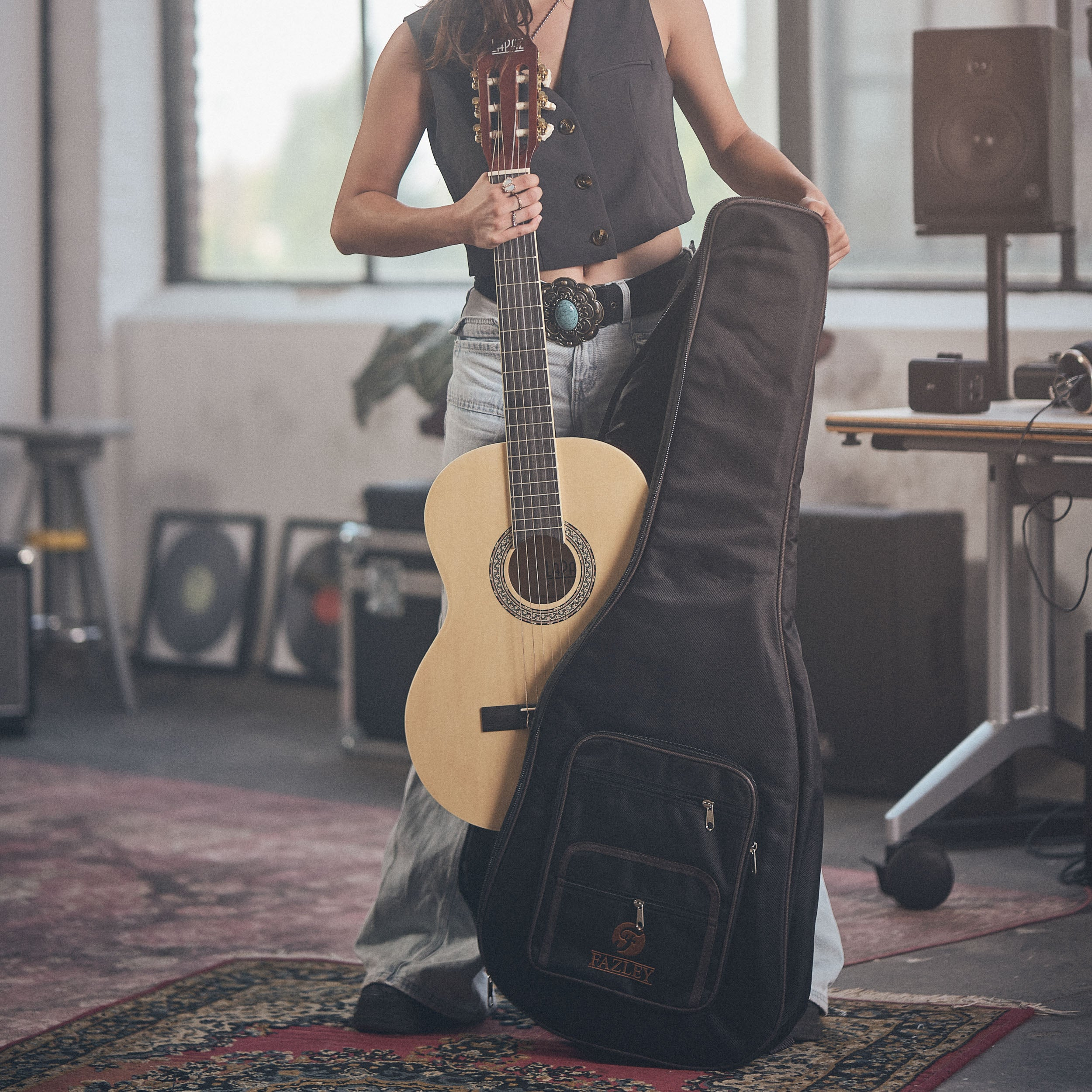 Een vrouw houdt een gitaar en tas vast in een kamer met vloerkleed en studioapparatuur.