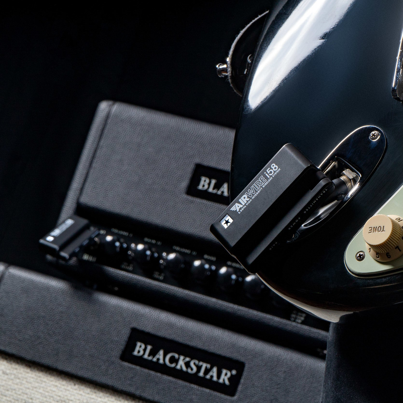 A black guitar transmitter is plugged into a black electric guitar, with guitar amps beside it.