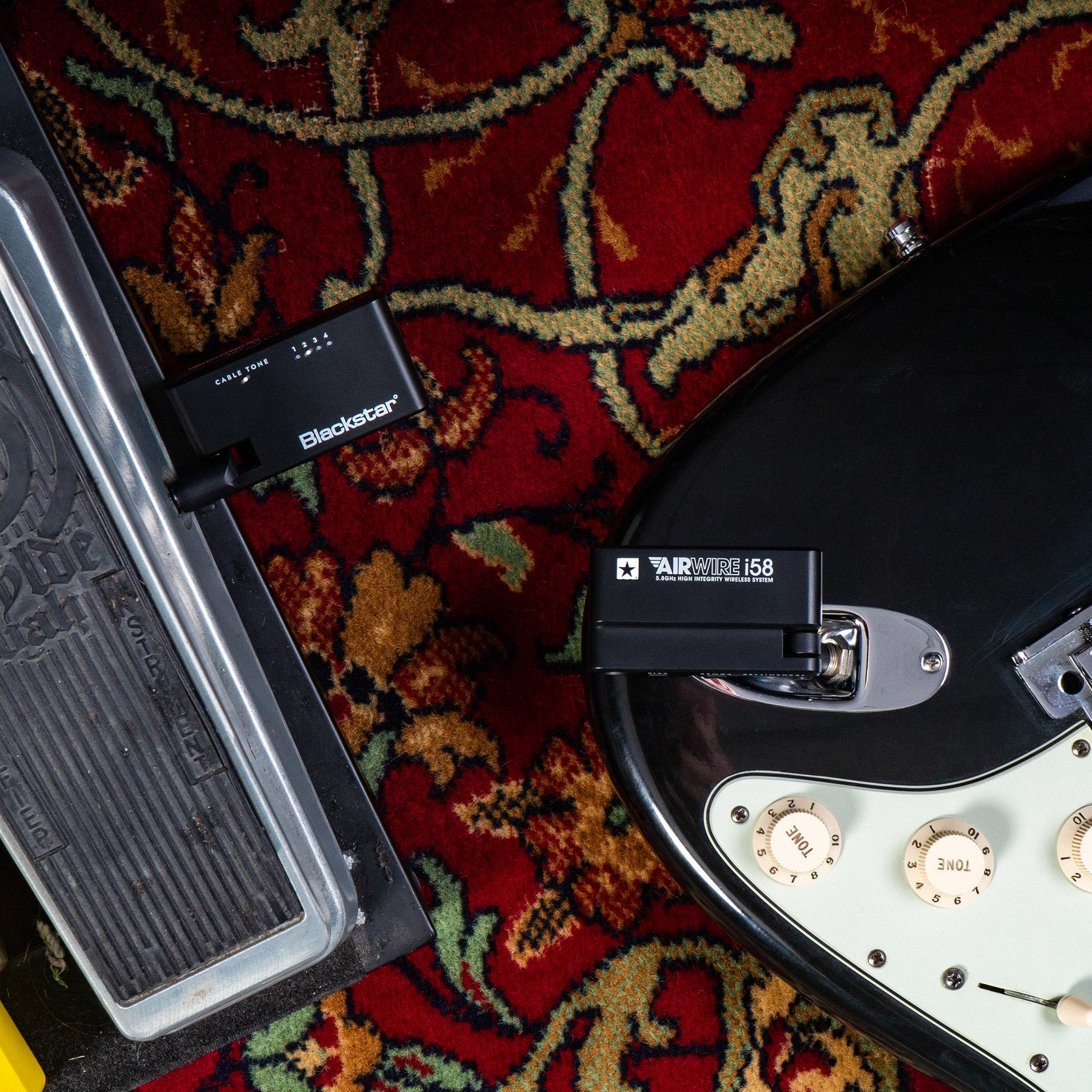 A black electric guitar transmitter and a pedal lie on a red rug.