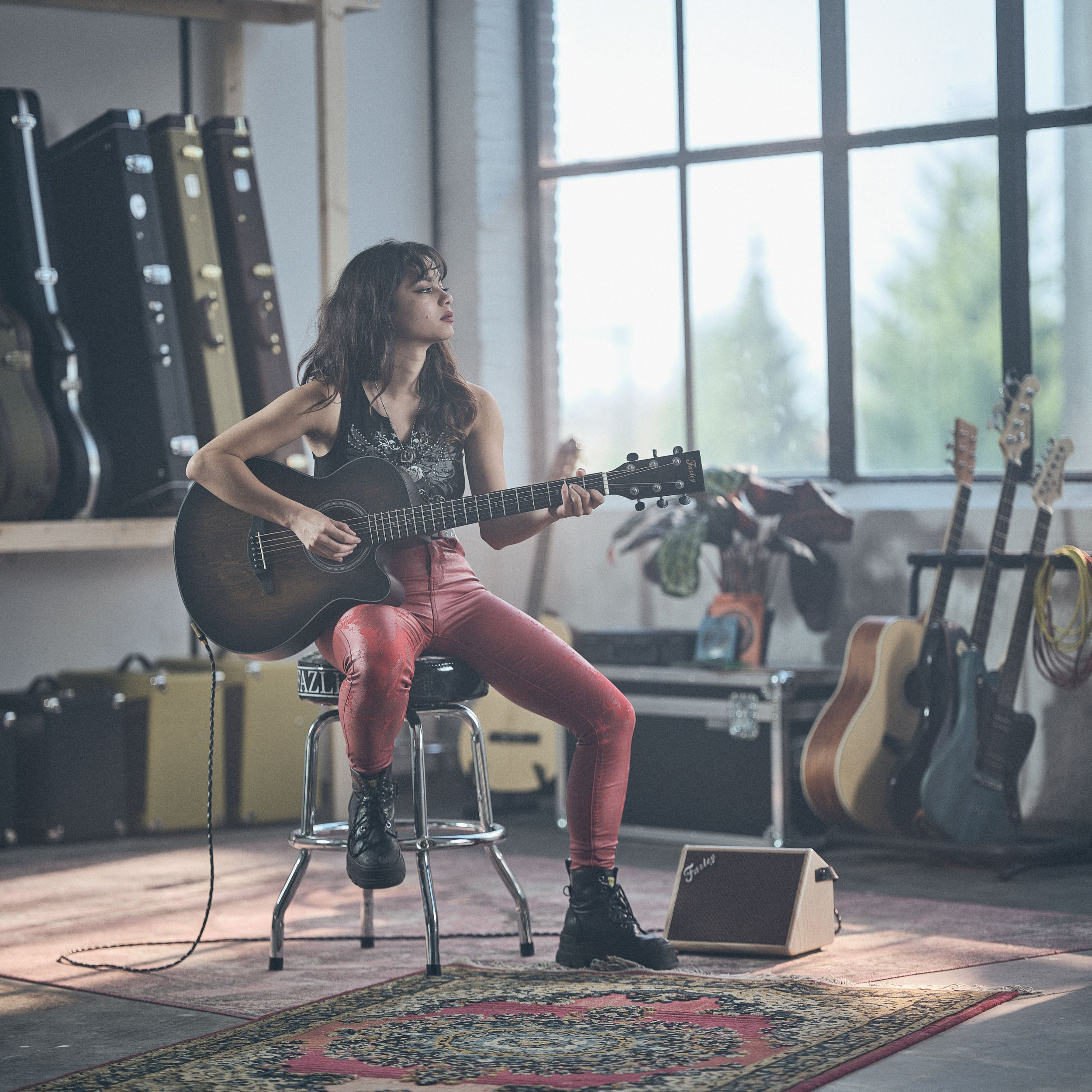 Vrouw speelt akoestische gitaar, zit op kruk, rode leren broek, gitaarkoffers op achtergrond.