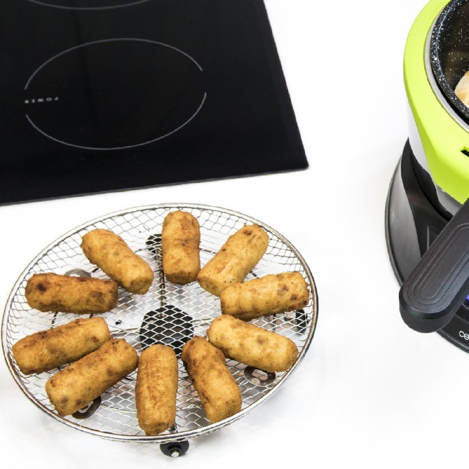 Croquetas fritas en una rejilla redonda de metal junto a una placa de cocina y una freidora.
