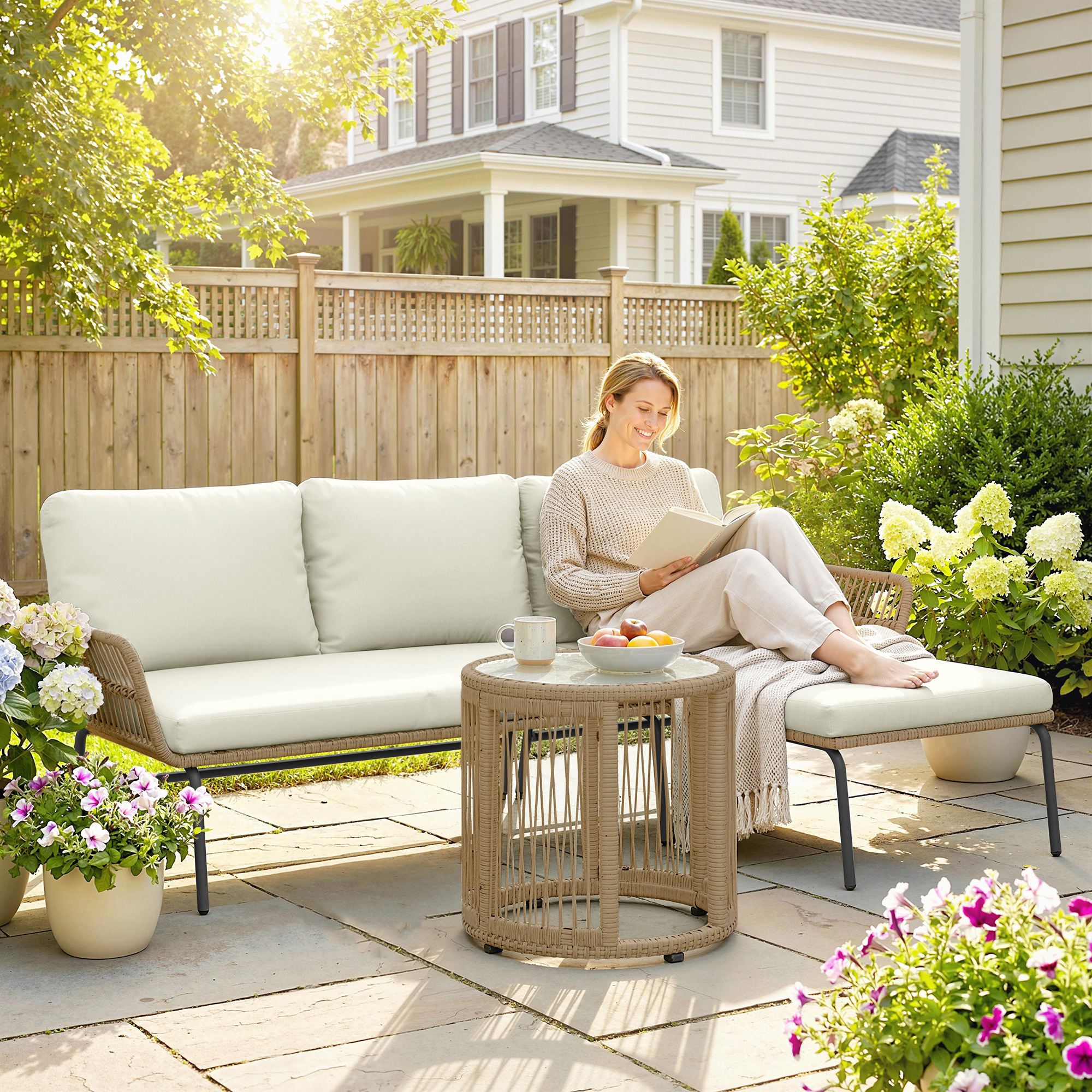 Una mujer lee un libro en un sofá de patio junto a una mesa auxiliar, con flores.