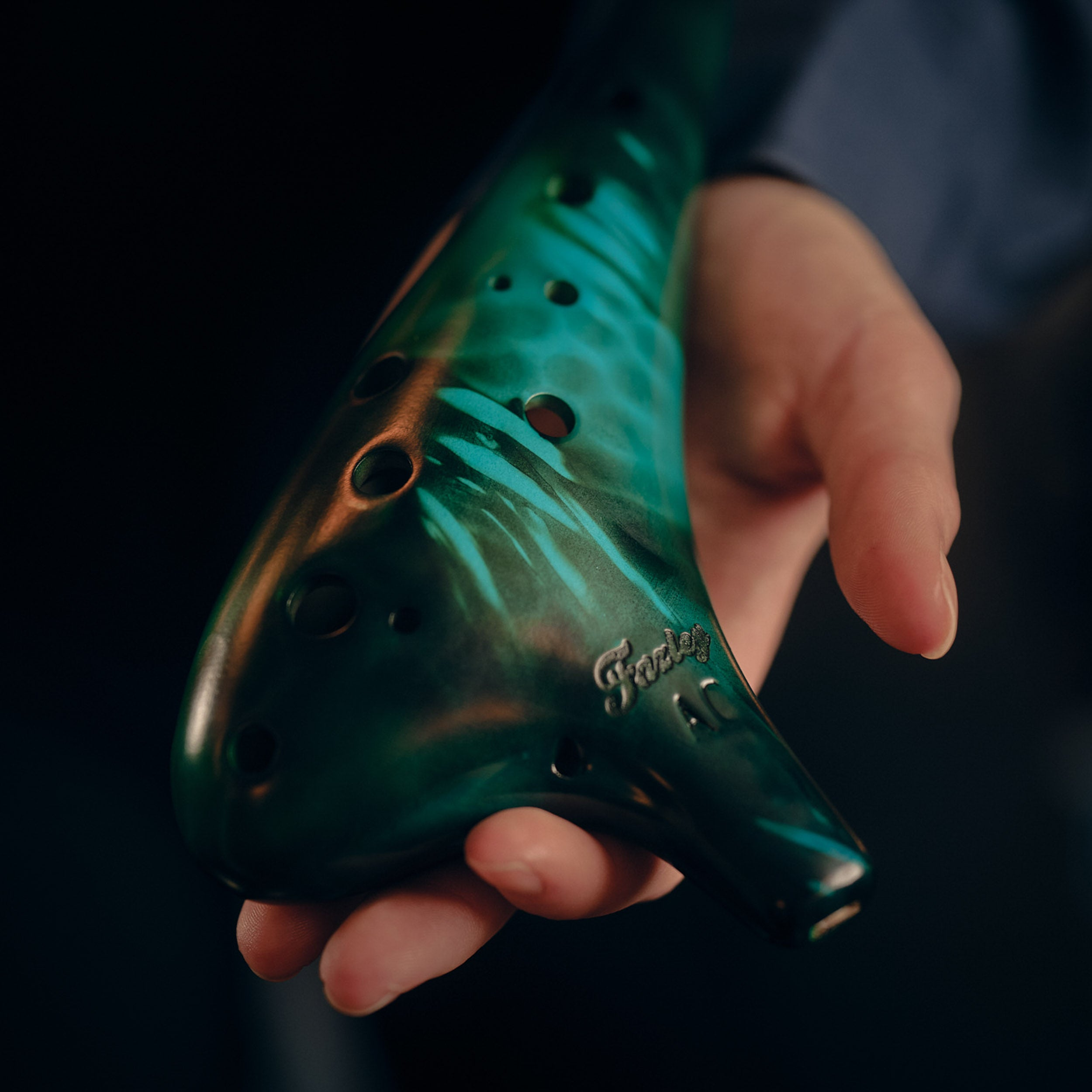A hand holds a green ocarina with black swirls. Dark background.