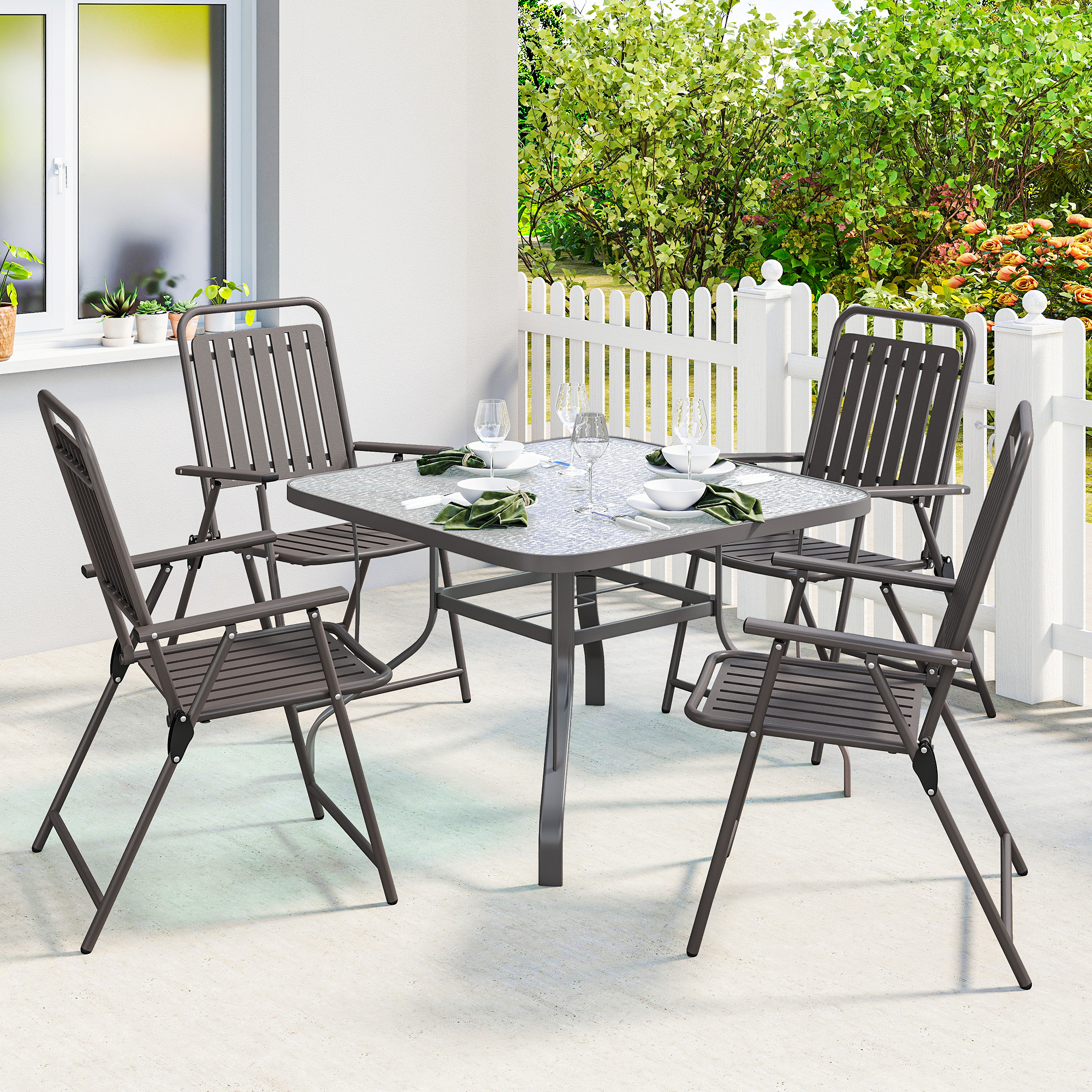 Mesa de cristal con sillas grises al aire libre, junto a valla blanca y plantas verdes.