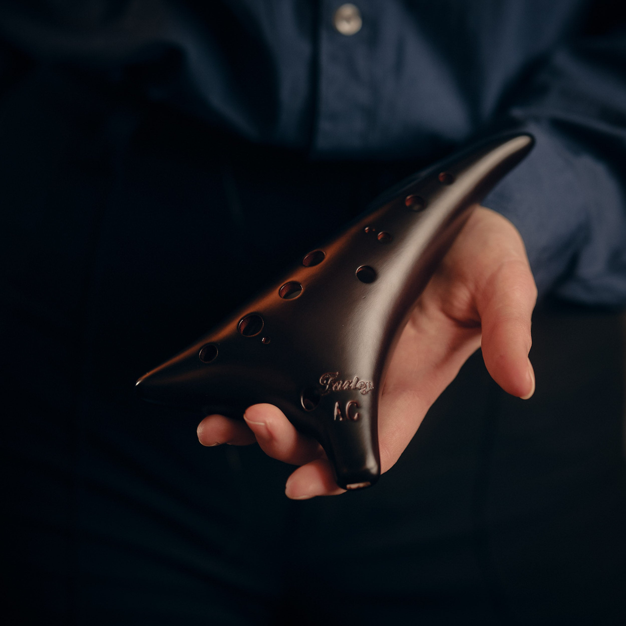 A hand holds a dark ocarina with many holes and 'Fletch A.C.' inscription.