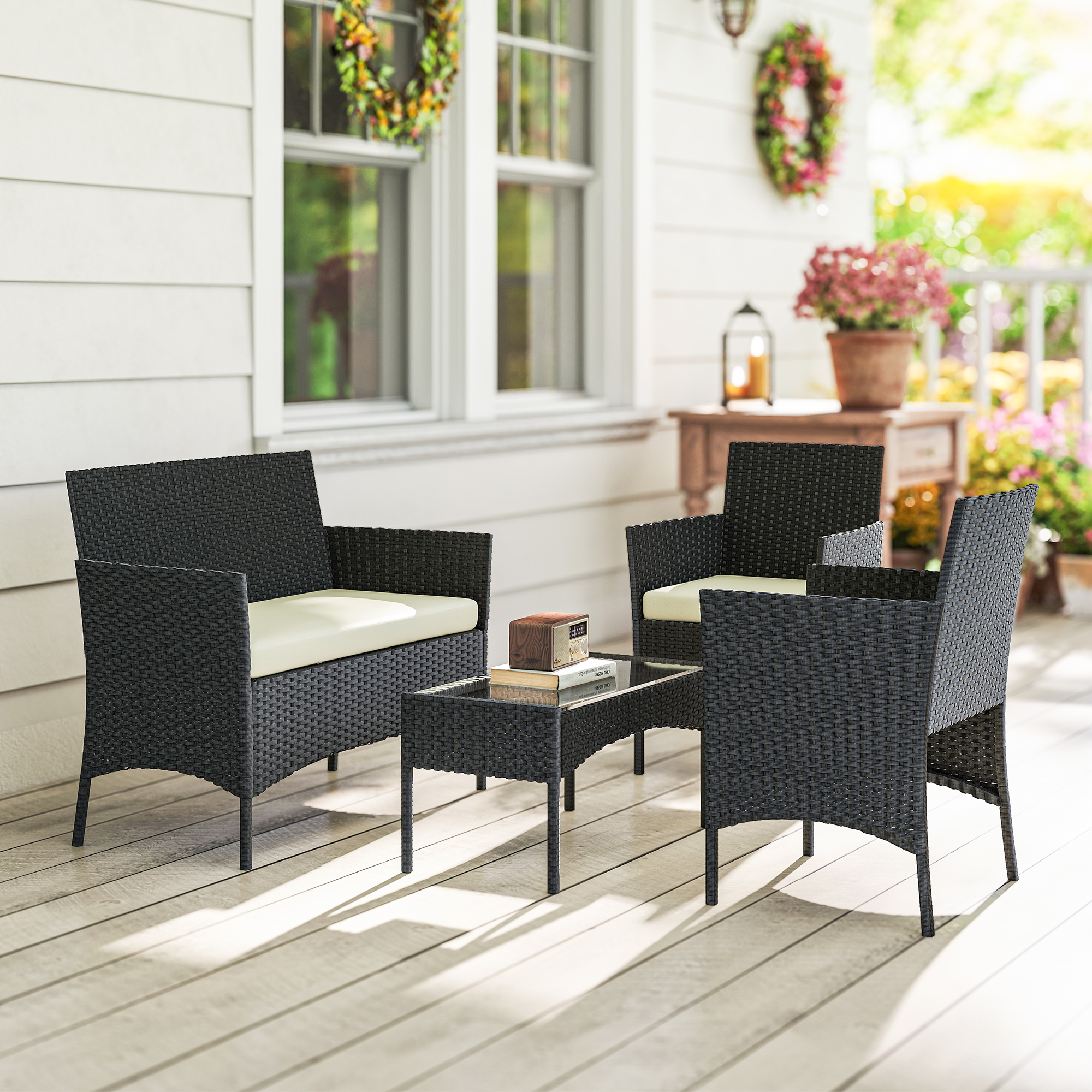 Muebles negros de ratán con cojines color crema en un porche de madera con flores y sol.