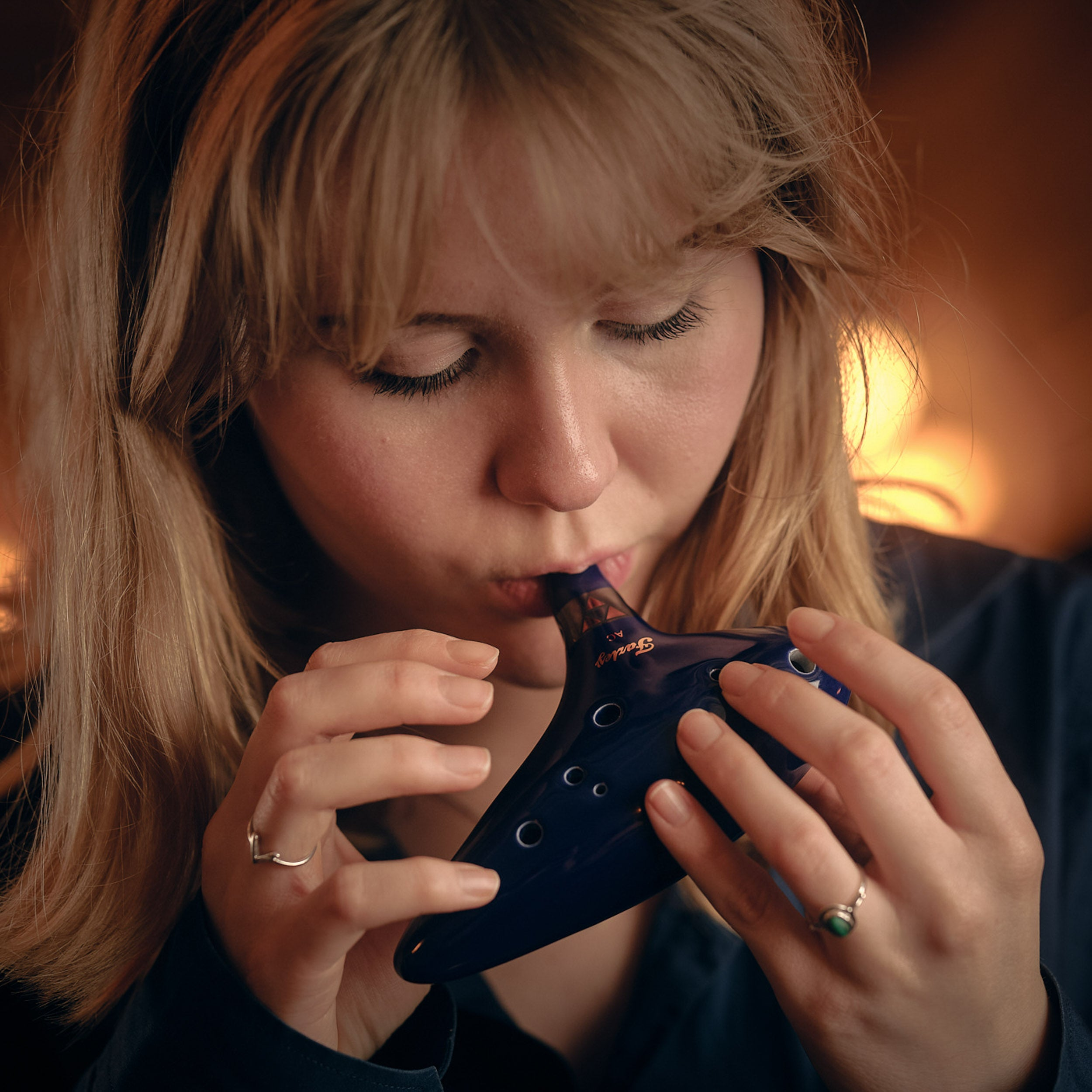 A woman with blonde hair plays a dark blue ocarina. She is wearing rings.