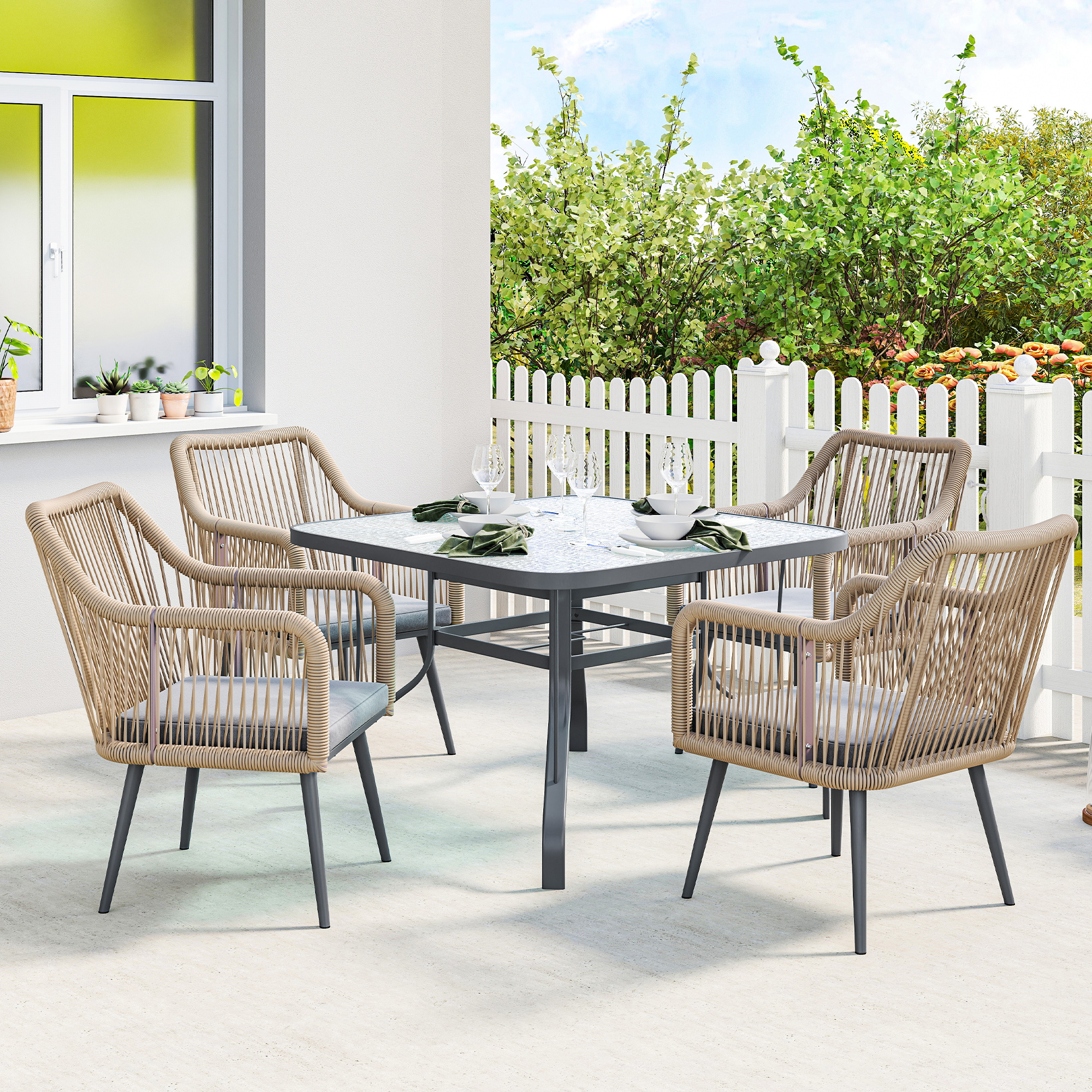 Una mesa de comedor con cuatro sillas de ratán con cojines en un patio de cemento, con valla y árboles al fondo.