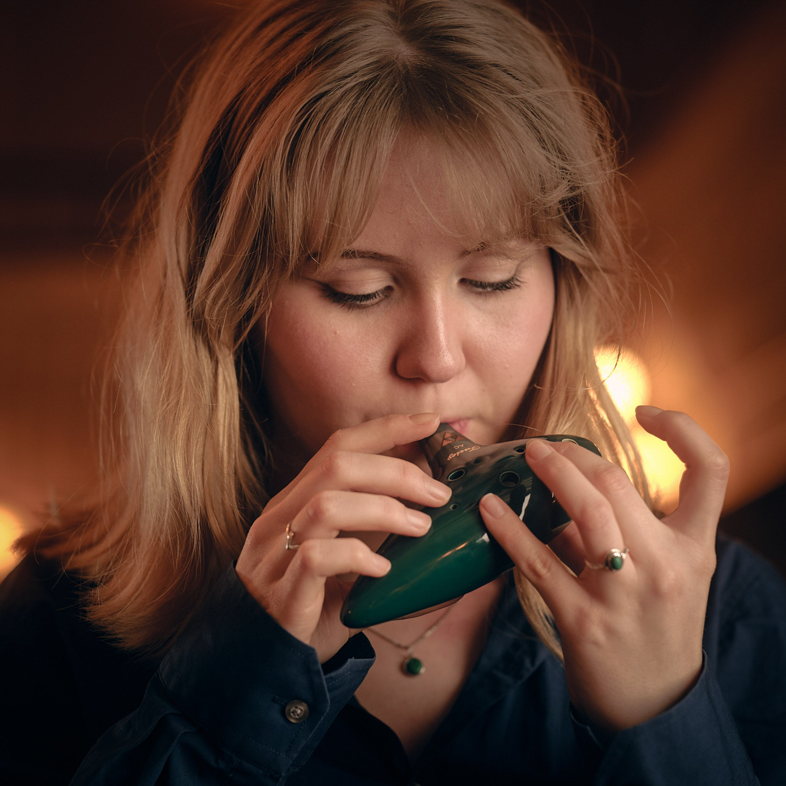 A young woman with blonde hair plays a green ocarina.