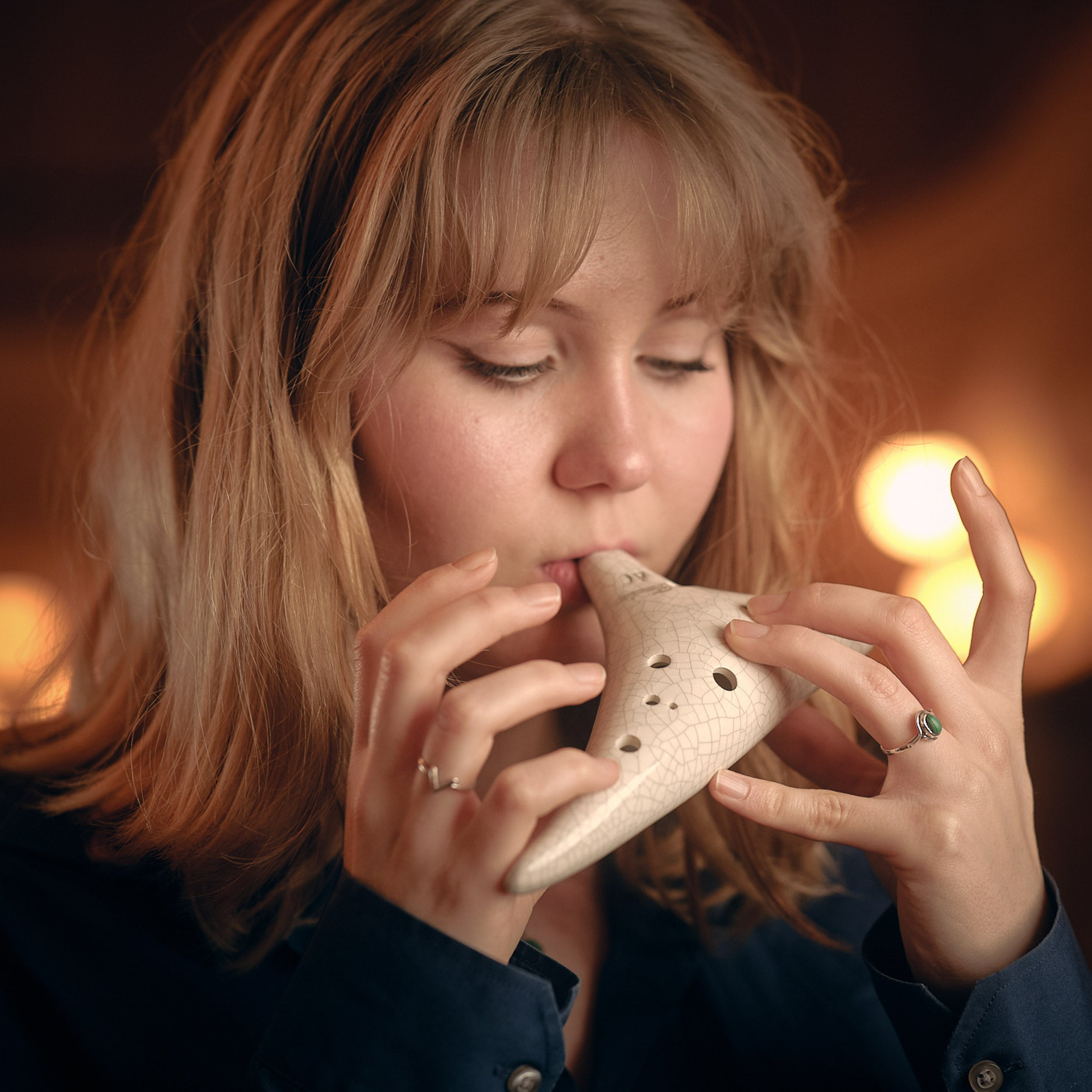 A blonde woman plays a white ocarina, with blurred lights in the background.