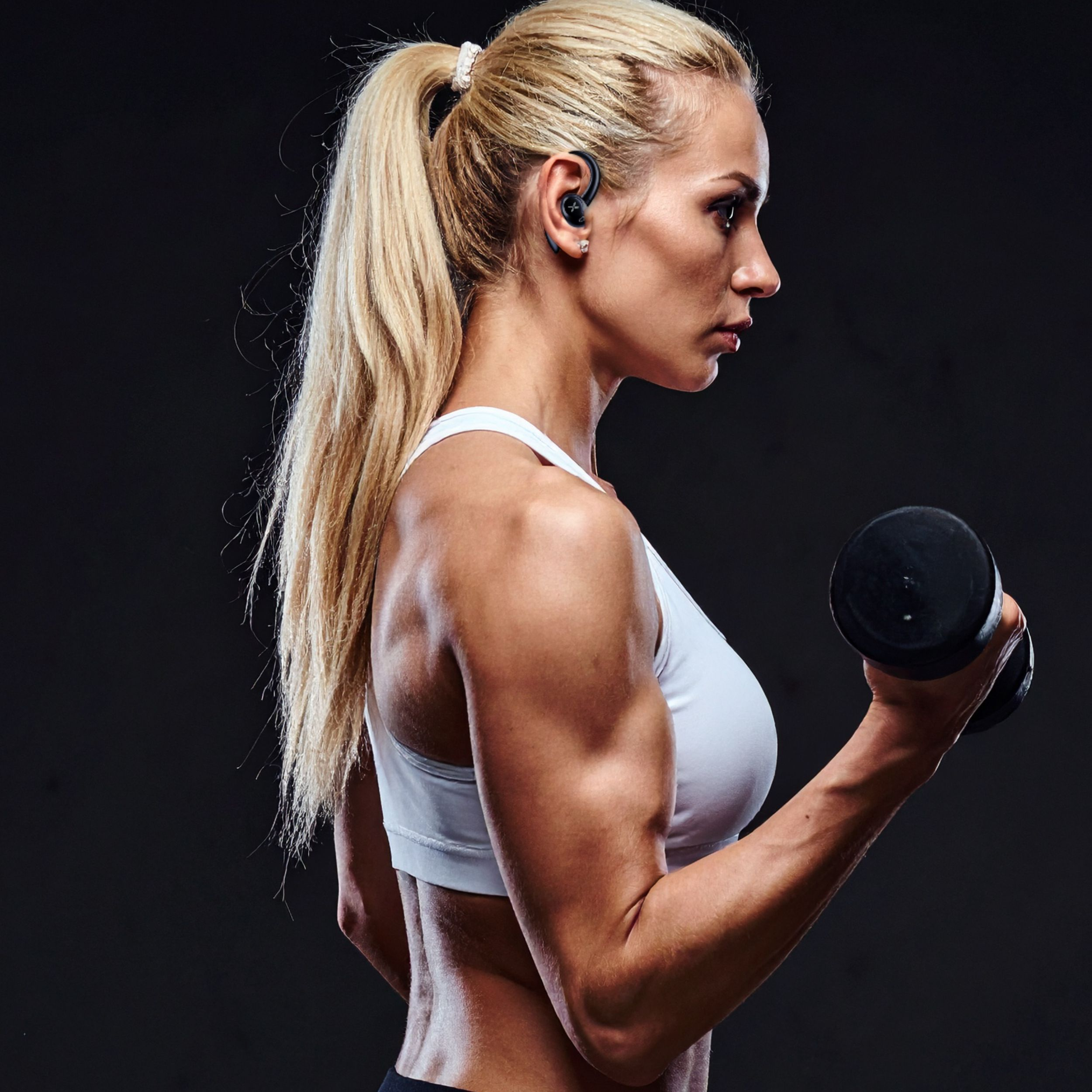 Kobieta z blond kucykiem podnosi hantle, ubrana w biały strój sportowy.