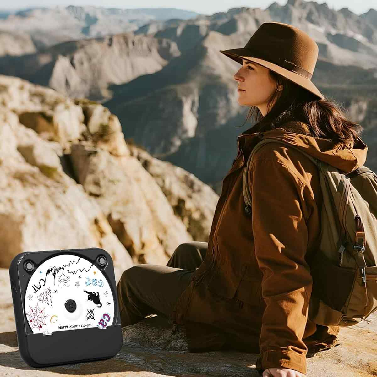 Eine Frau in brauner Kleidung sitzt auf einem Felsen mit einem CD-Player und Bergen im Hintergrund.