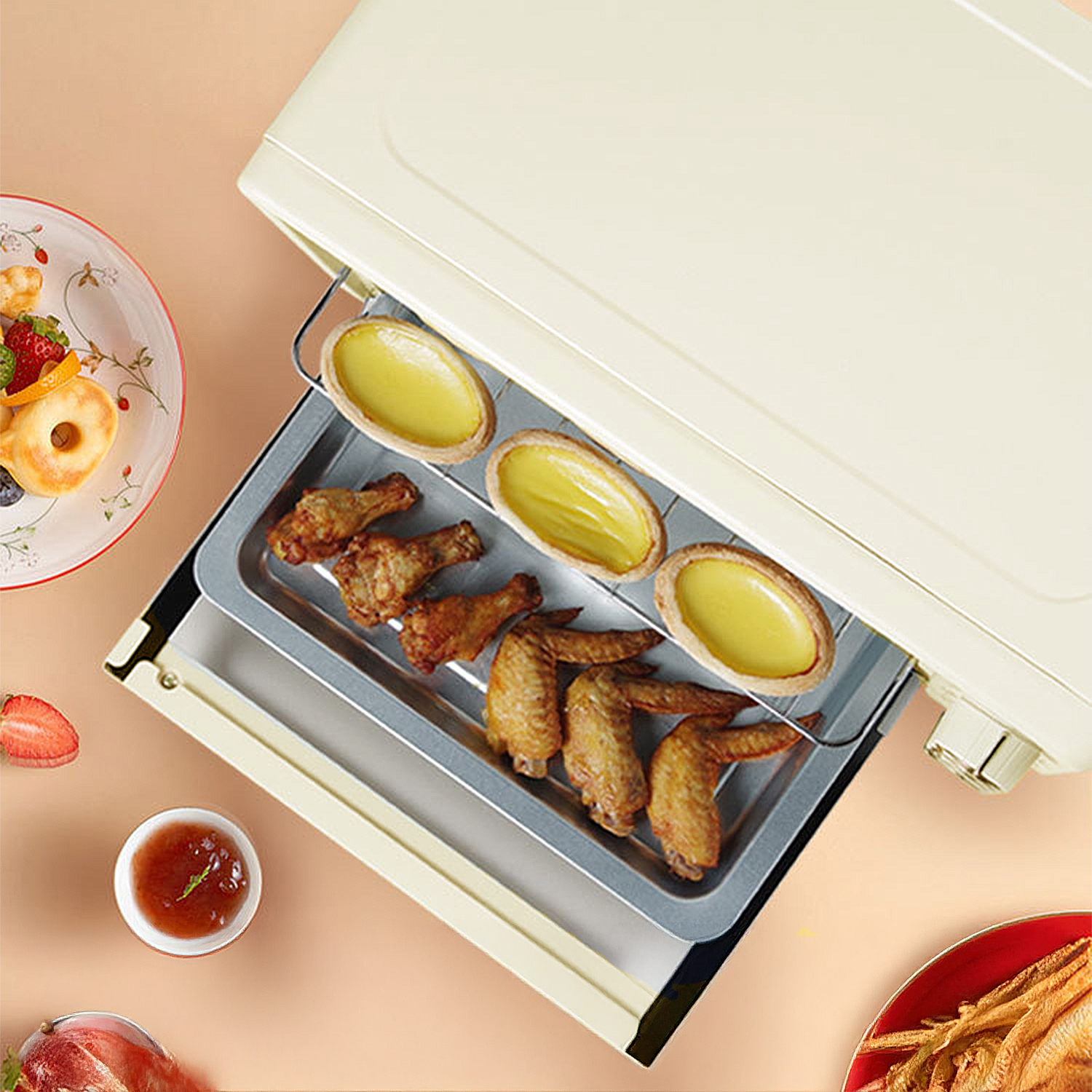 Ein Toaster mit Hähnchenflügeln und Eiertörtchen innen, daneben Obst und Sauce.