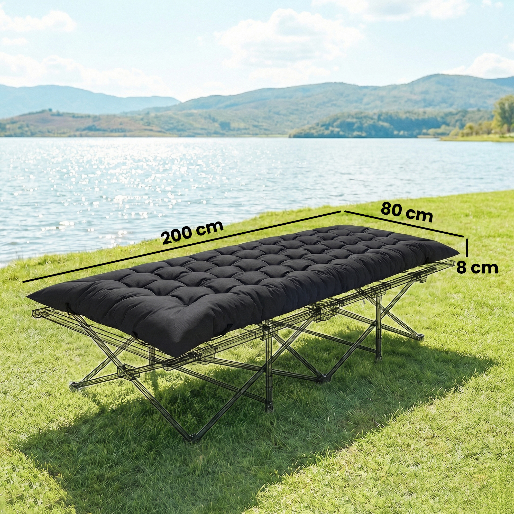 Cama plegable negra con colchón oscuro sobre hierba verde junto a un lago.