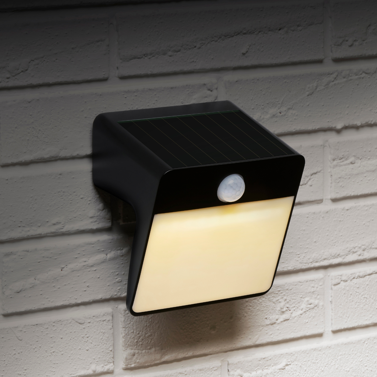 A black solar-powered outdoor light with a motion sensor is mounted on a white brick wall.