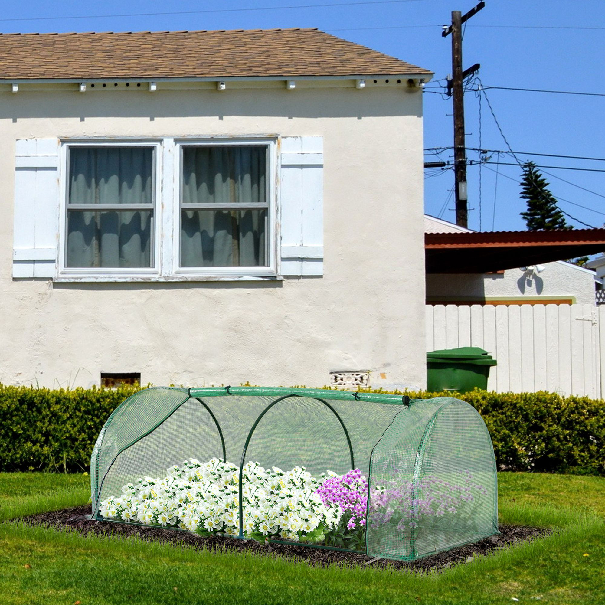 Un mini-invernadero verde con flores blancas y moradas dentro, frente a una casa.