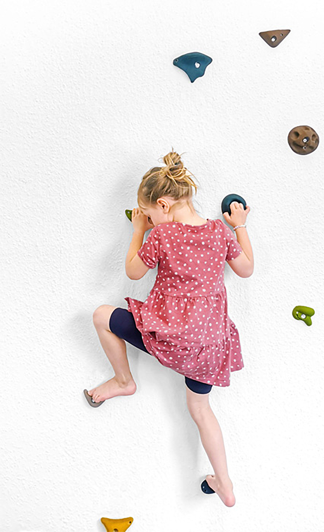 Ein kleines Mädchen klettert an einer weißen Wand mit bunten Griffen.