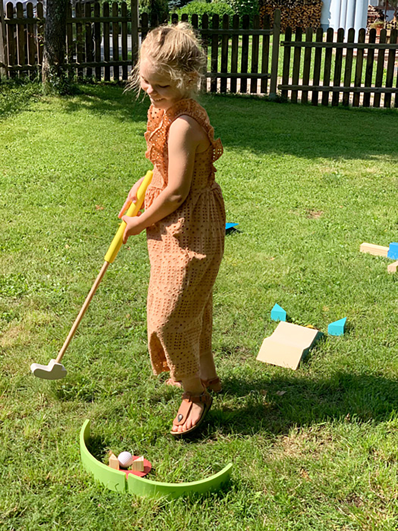 Ein kleines Mädchen in einem orangefarbenen Kleid spielt Minigolf im Garten.