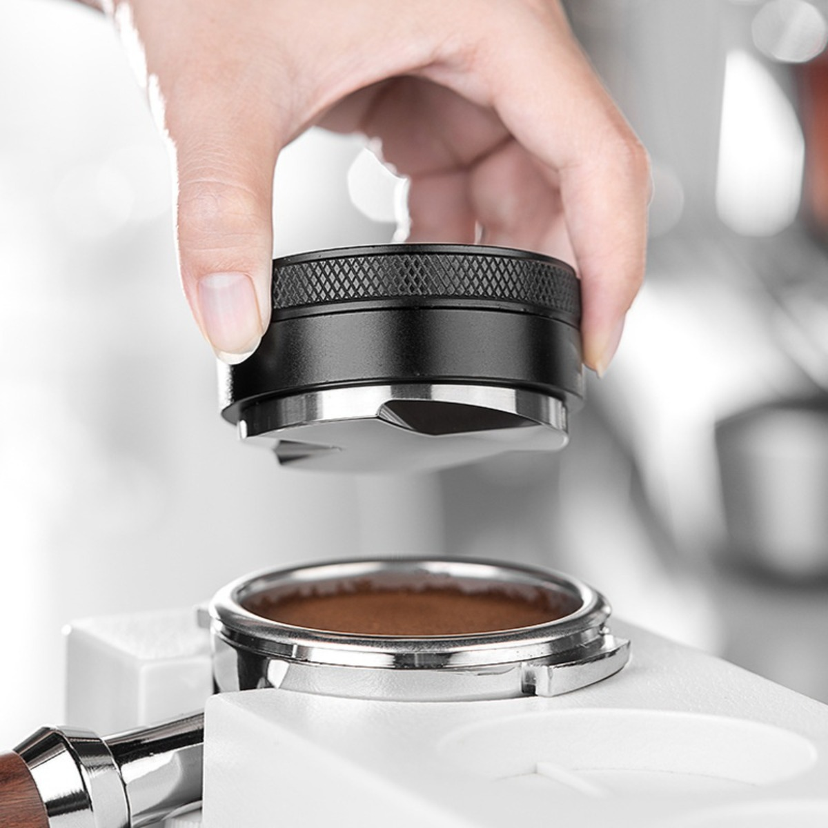 Una mano tiene un tamper nero per macchina da caffè sopra un portafiltro con caffè macinato.