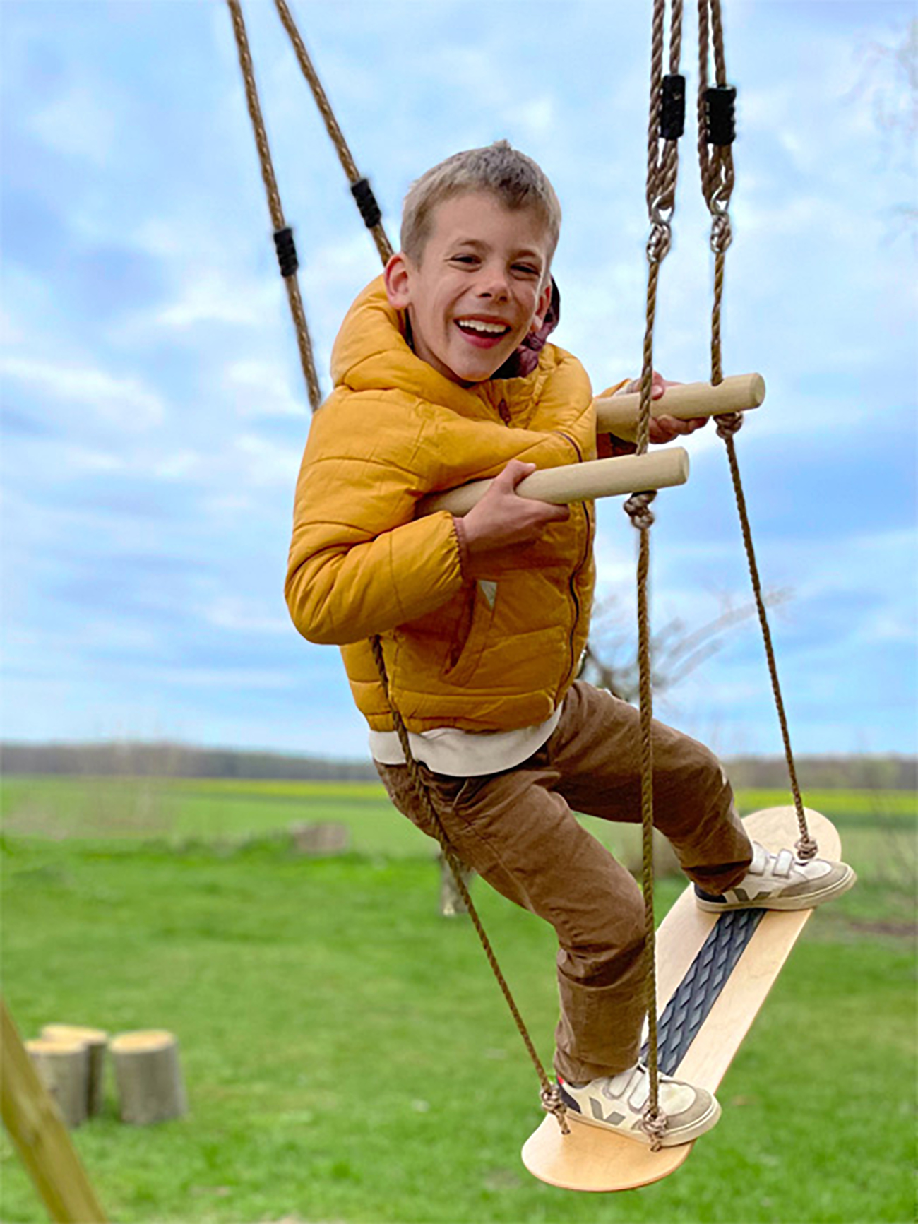 Ein Junge in einer gelben Jacke lacht, während er auf einer Schaukel mit Holzgriffen und einem Skateboard-Sitz schwingt.