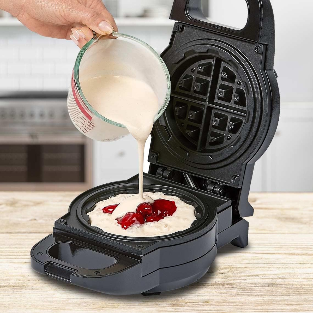 Ein Glas mit Teig wird über eine Waffelform gegossen, in der sich Kirschen befinden.