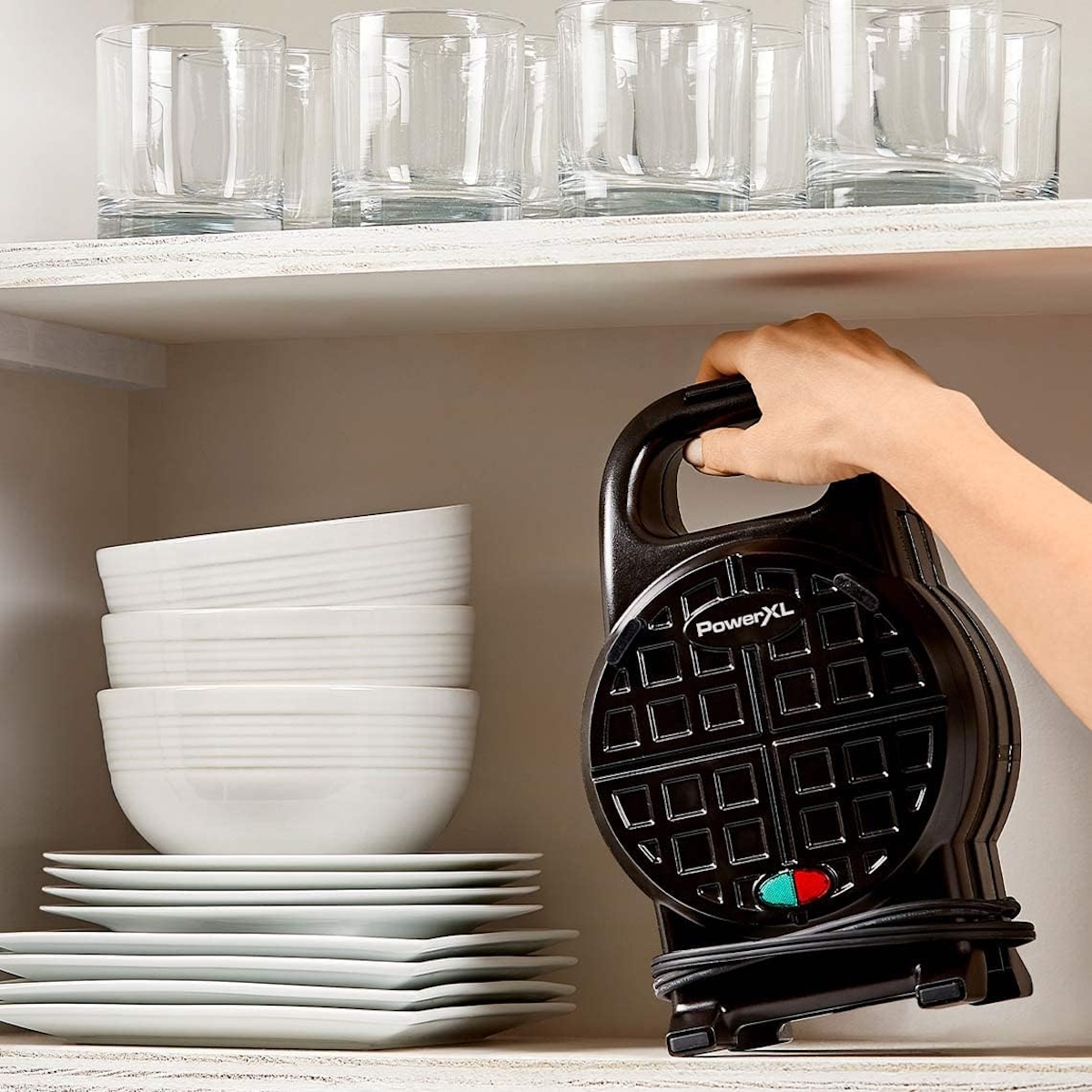Eine Hand hält einen schwarzen Waffelmaker im Regal mit gestapelten weißen Schüsseln und Tellern.