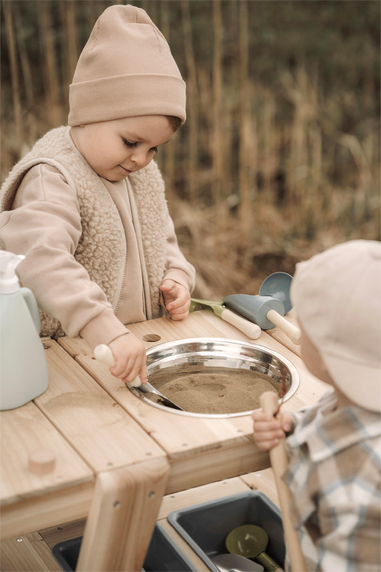 Zwei Kinder spielen mit Sand an einem Holztisch im Freien.