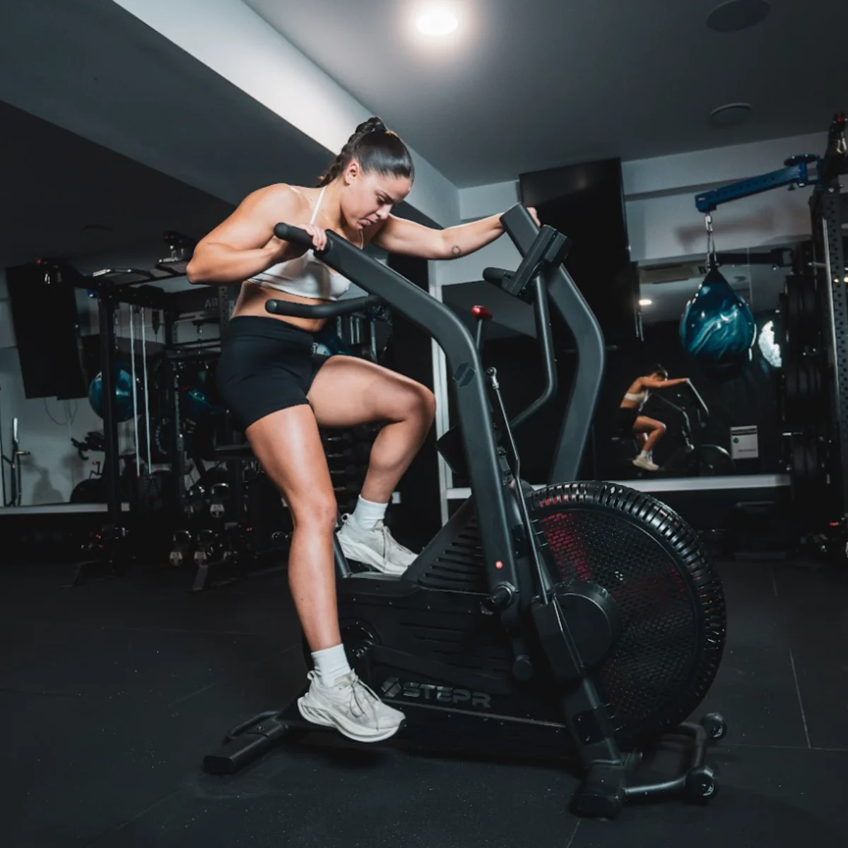 Vrouw op hometrainer in sportschool. Ze draagt witte sportbeha en zwarte korte broek.